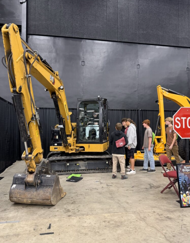 A group of children and adults viewing a yellow excavator at an indoor exhibit, with a red stop sign and a black wall in the background.