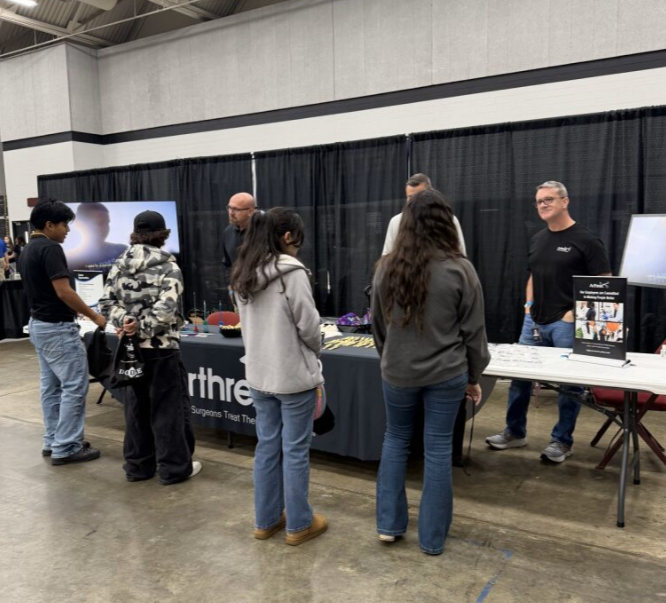 People visiting an exhibition booth at a convention or trade show, with merchandise displayed on tables and a large screen in the background.