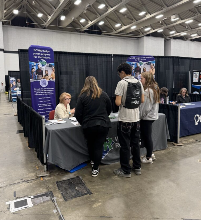 A group of five people at an expo or career fair booth with a black tablecloth and informational banners, in a large indoor space with high ceilings.