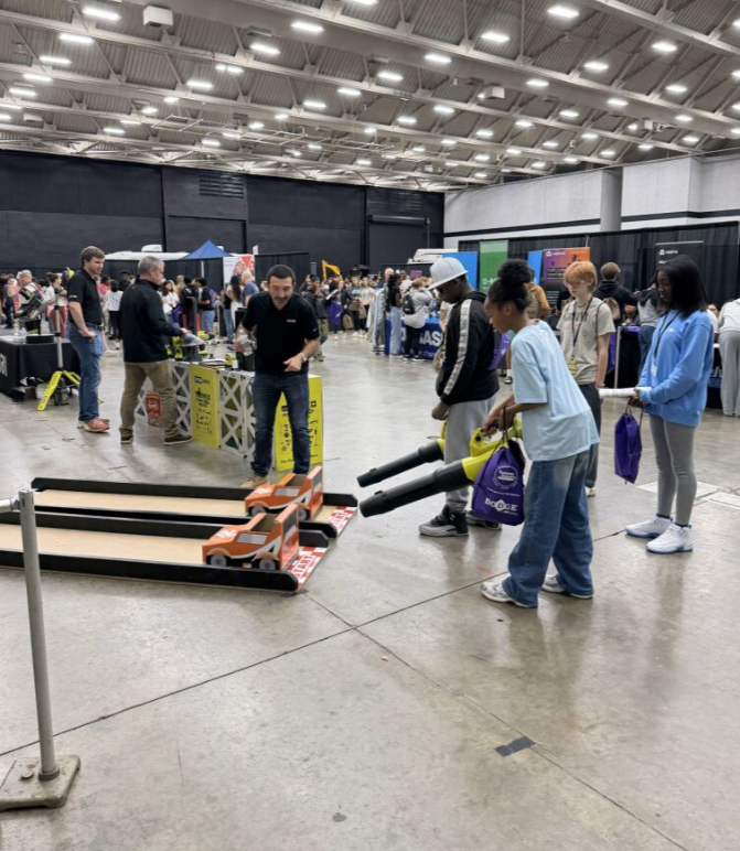 People at a tech or science convention engaging with interactive exhibits, including a mini race car track and a cannon-like device.