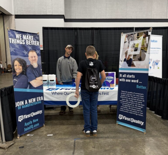 A young man and a woman with a backpack standing at a trade show booth talking to a representative. The booth displays banners for First Quality, a manufacturing company.