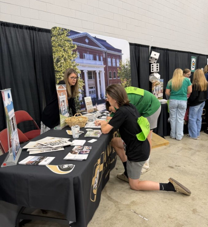 Person kneeling at informational booth at indoor event, interacting with woman behind table, with large building backdrop and other attendees nearby.