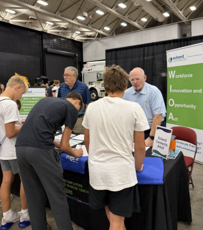 People at a booth during a career or job fair, with a banner promoting Eckerd Connects Workforce, Innovation, and Opportunity, and a truck visible in the background.