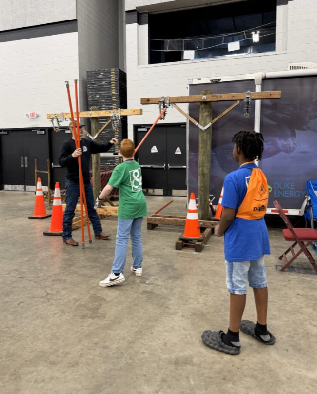 Three children observing a construction or assembly activity with two adults inside a spacious indoor facility. The scene includes orange safety cones, wooden structures, and safety equipment.