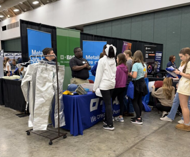Group of young people at a technology booth in an exhibition hall, engaging with a representative and exploring displayed items.