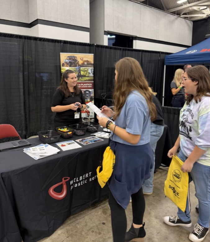 Two young women talking at a trade booth about Wilbert plastic service, with a woman behind the booth smiling and holding a pen. The booth has promotional materials and a black tablecloth with the Wilbert logo. Other people are visible in the backgro
