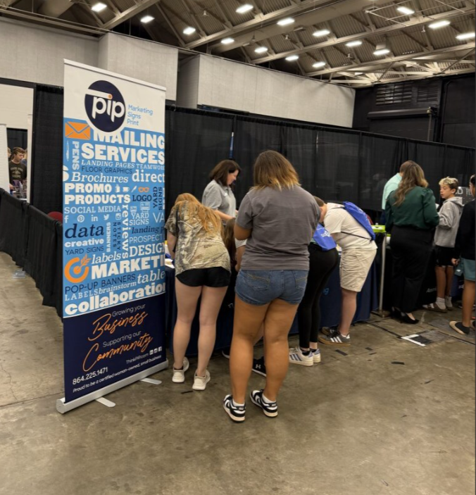 Several people browsing a booth at an event, with a tall blue and white sign that reads 'PIP Mailing Services' listing various services like marketing, printing, and signs.