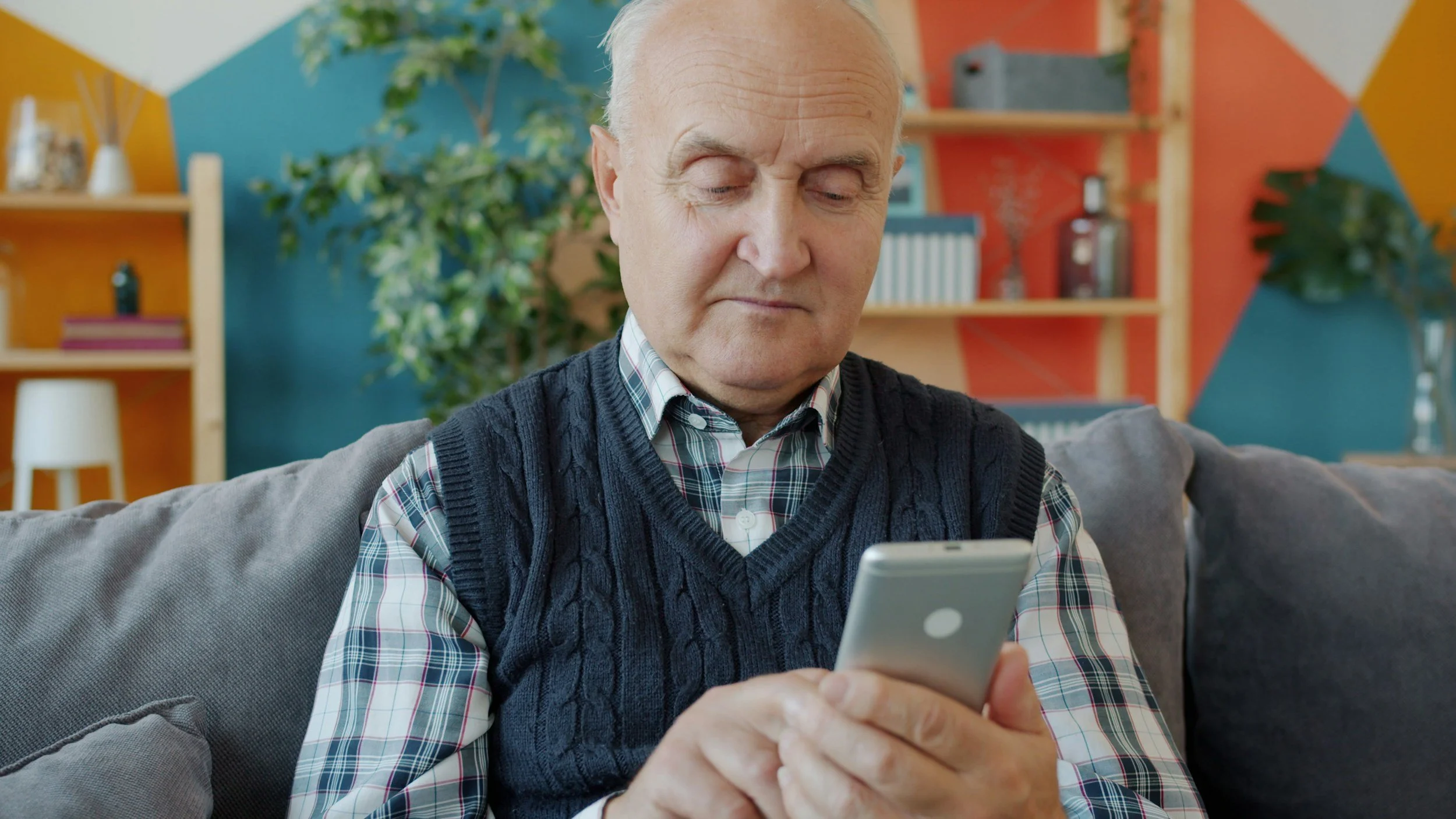 An elderly man with white hair, wearing a navy cable-knit sweater over a plaid shirt, sits on a gray couch and looks at his smartphone with a neutral expression. The background features colorful geometric wall art, shelves with various decor items, and a potted plant.