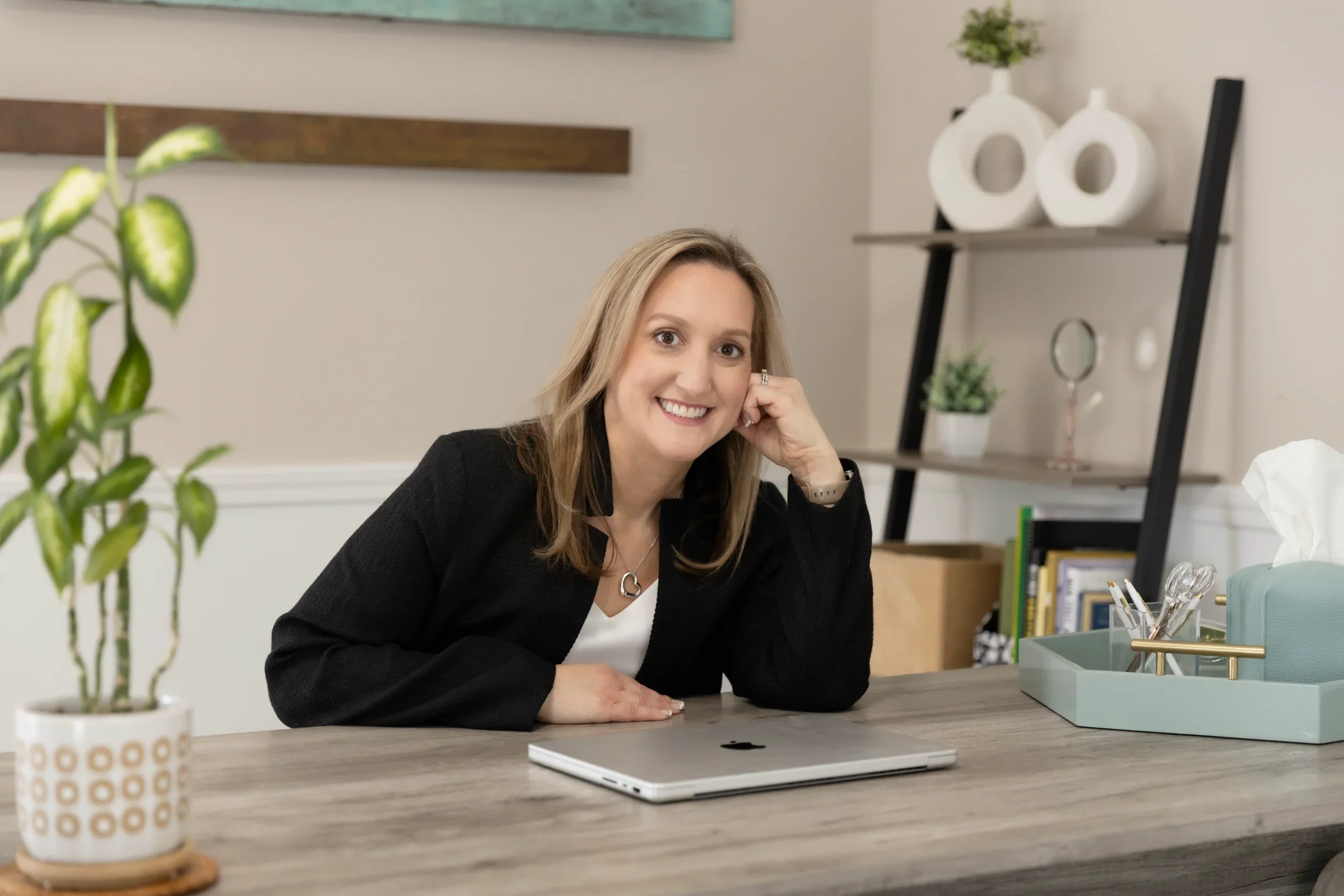 Tina sitting at her desk with a Macbook in front of her. She is wearing a black suit jacket and is leaned over with her head propped on her hand.