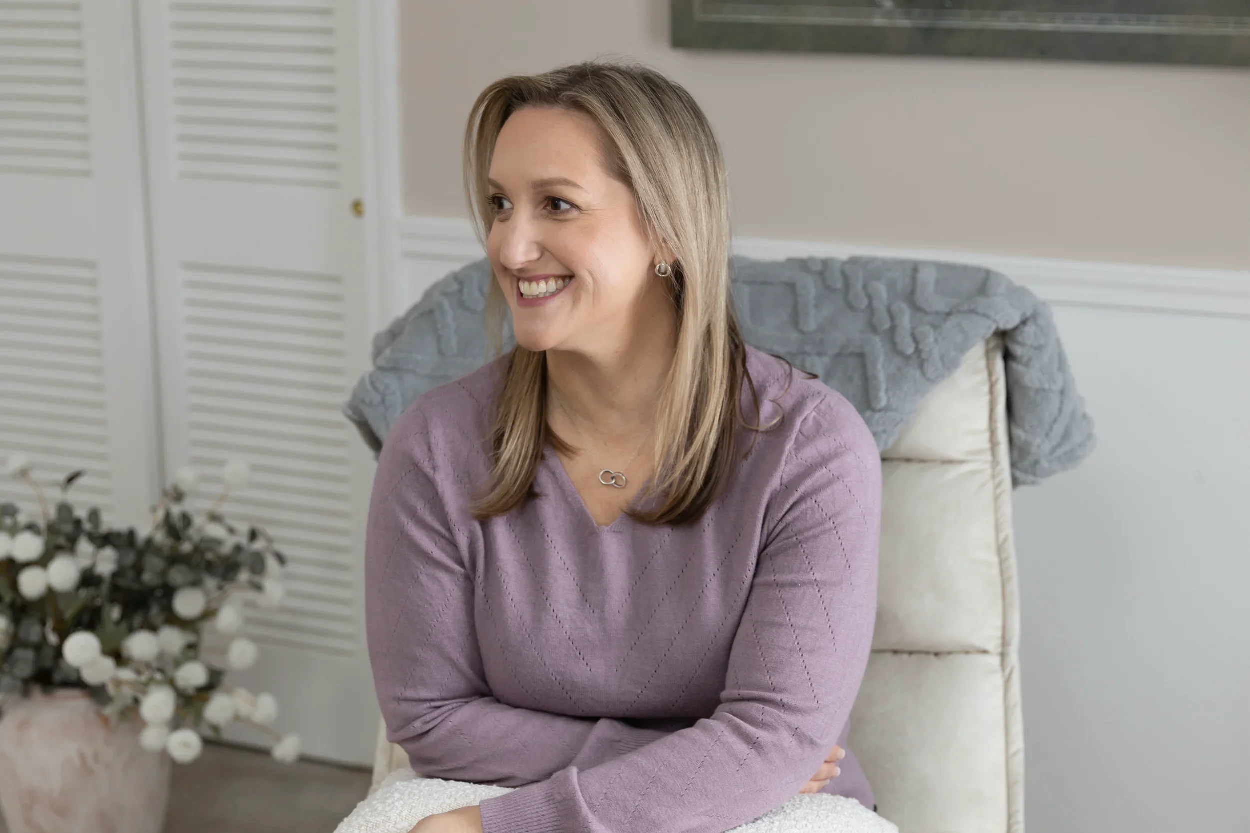 Tina is seated in a cream-colored chair wearing a purple sweater. She is looking off to the right and smiling.