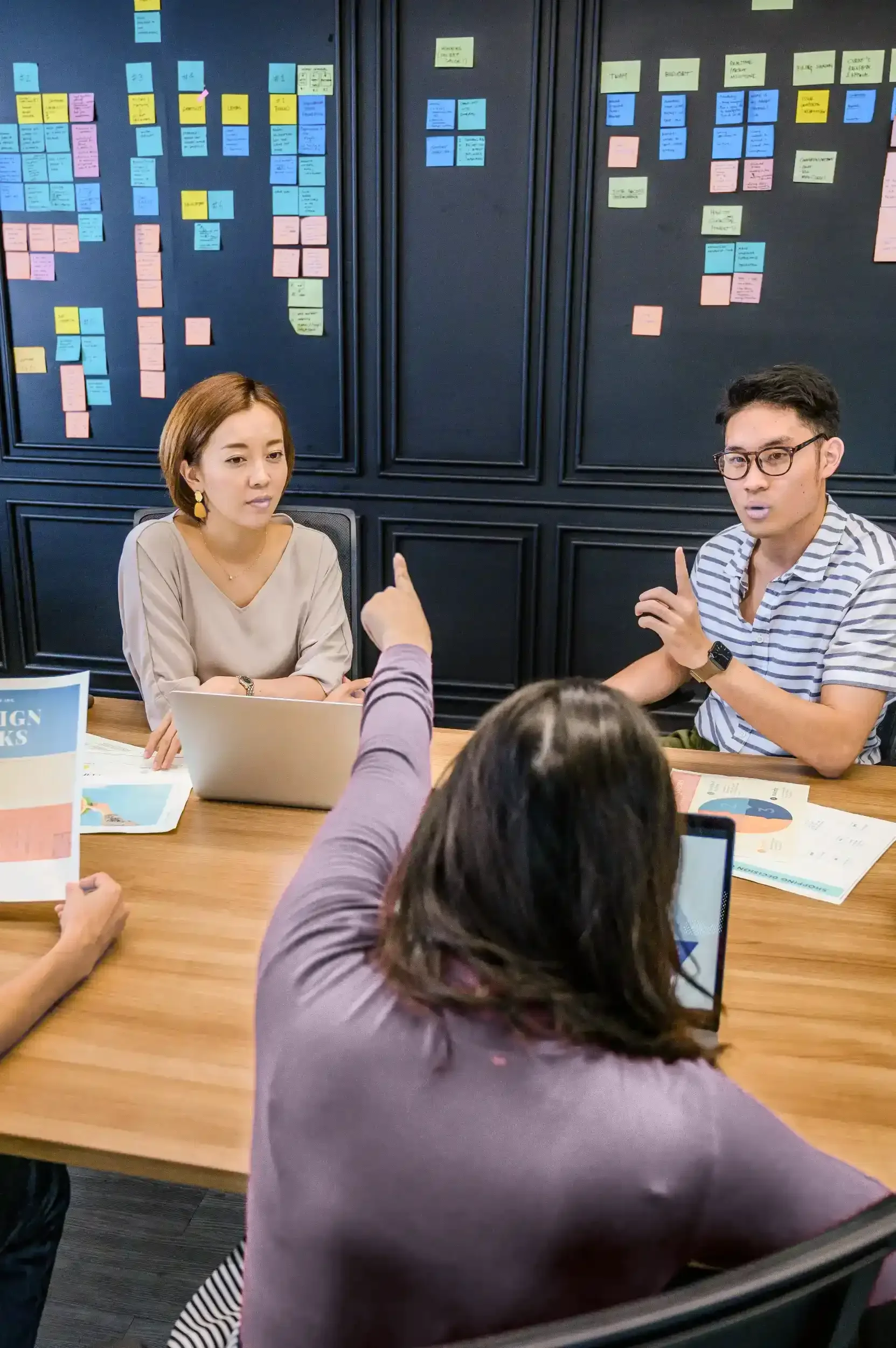A woman in a purple sweater is visible in the foreground, pointing at a wall full of post-it notes and talking to two others during a business meeting