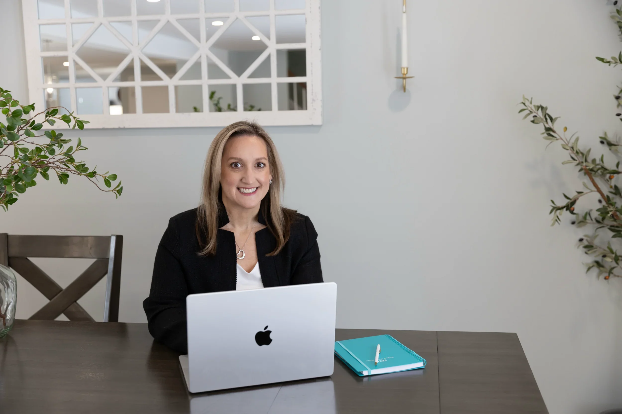 Tina sitting at her desk wearing a black blazer. In front of her is a Macbook laptop and a teal notebook.