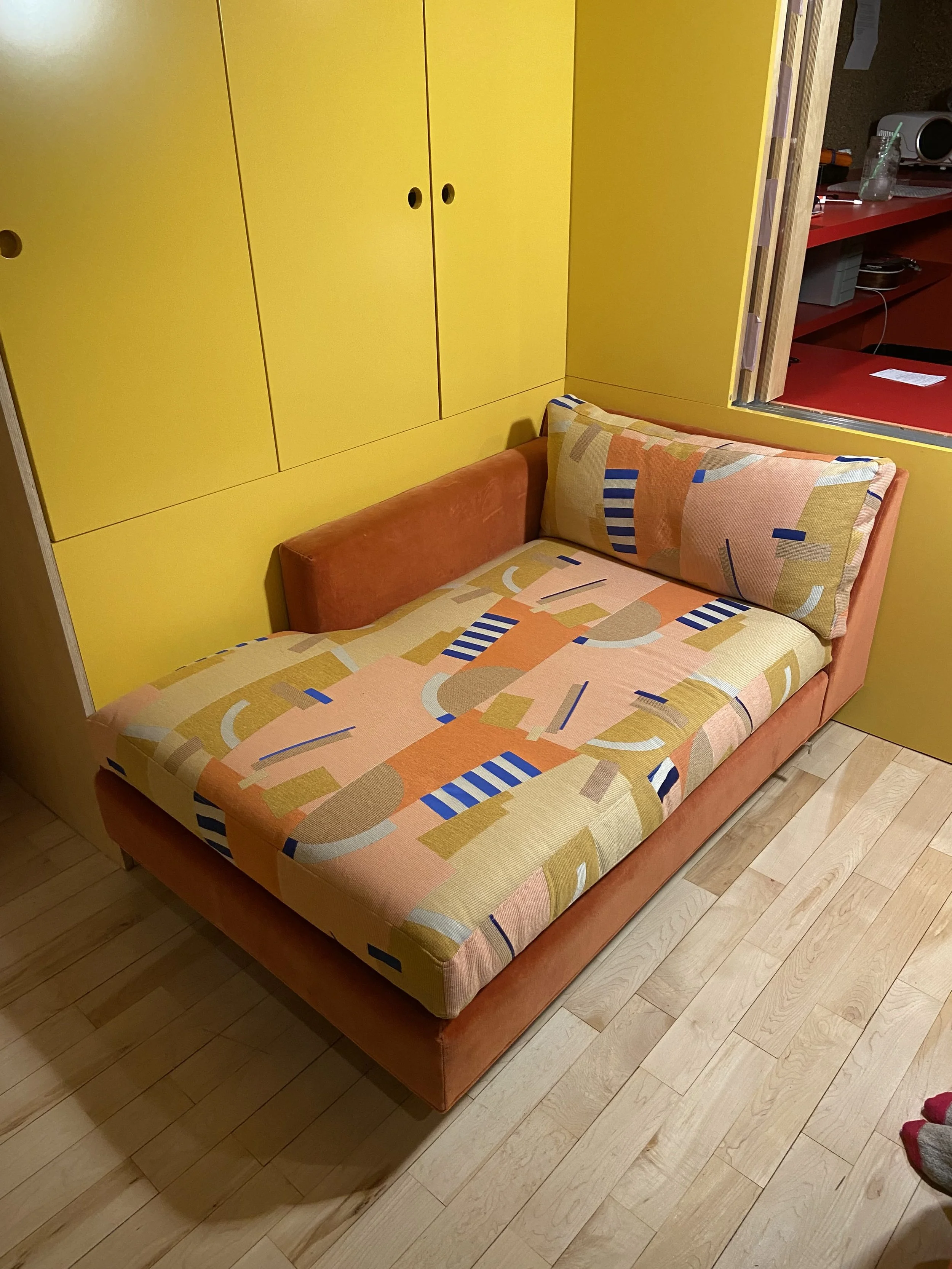 A colorful, abstract-patterned couch with a matching pillow, positioned against a yellow wall with built-in yellow cabinets.