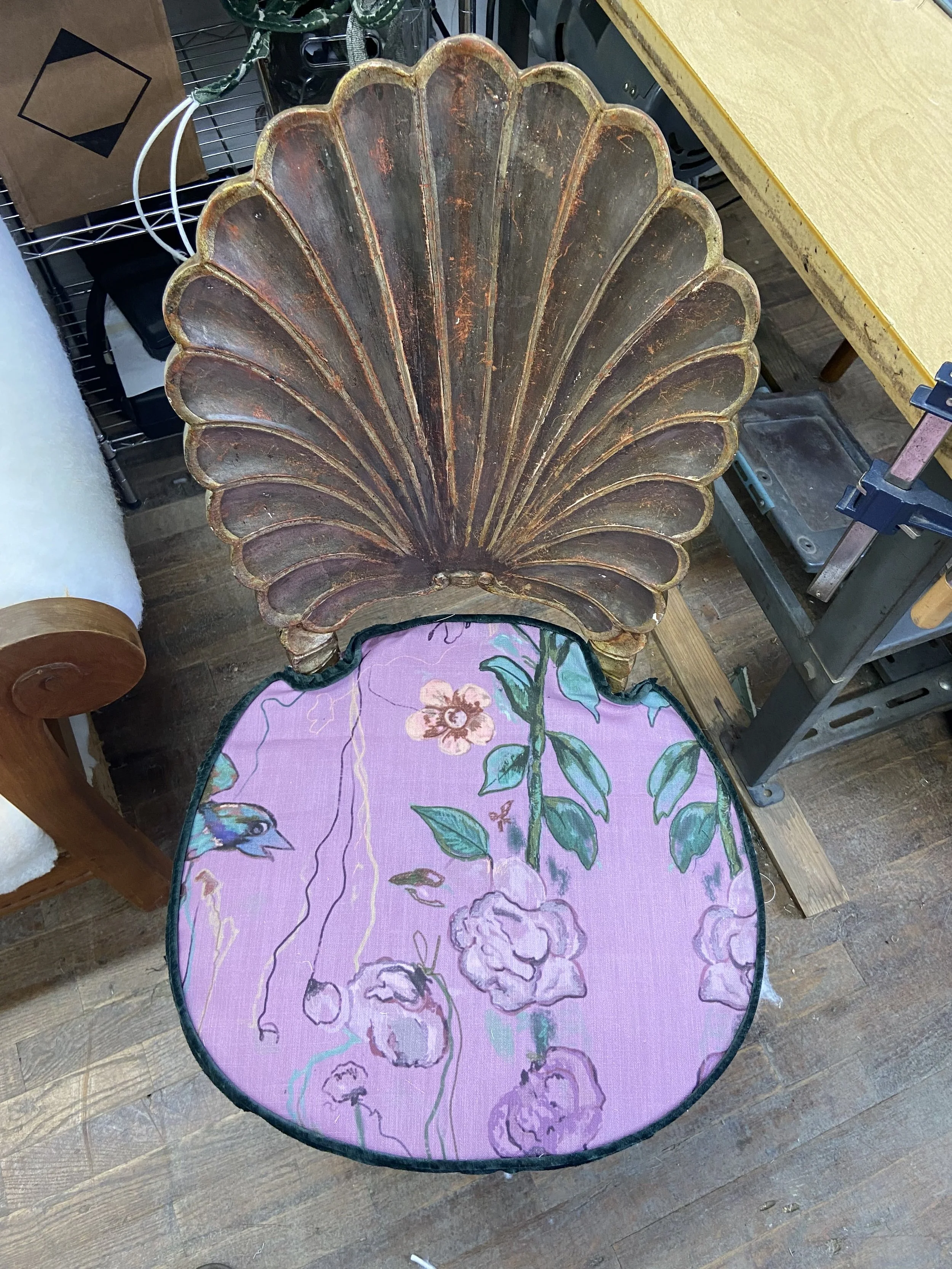 A vintage wooden chair with a seashell-shaped backrest and a pink cushioned seat featuring a floral pattern with flowers and leaves.