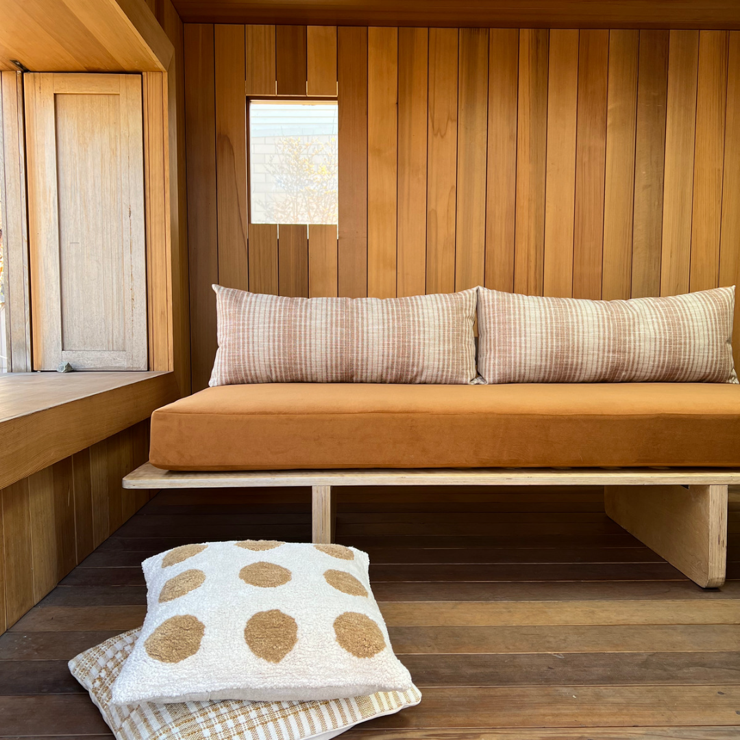 Interior of a wooden room with a cushioned bench, a large pillow, and a small polka-dotted pillow on the floor.
