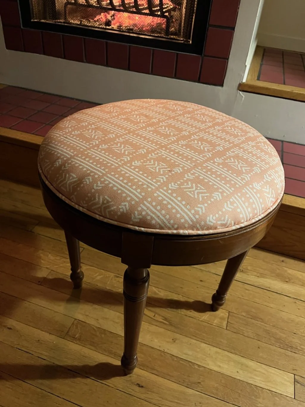 A wooden stool with a round, padded cushion covered in peach-colored fabric with a white geometric pattern, placed on hardwood floor in front of a brick fireplace.