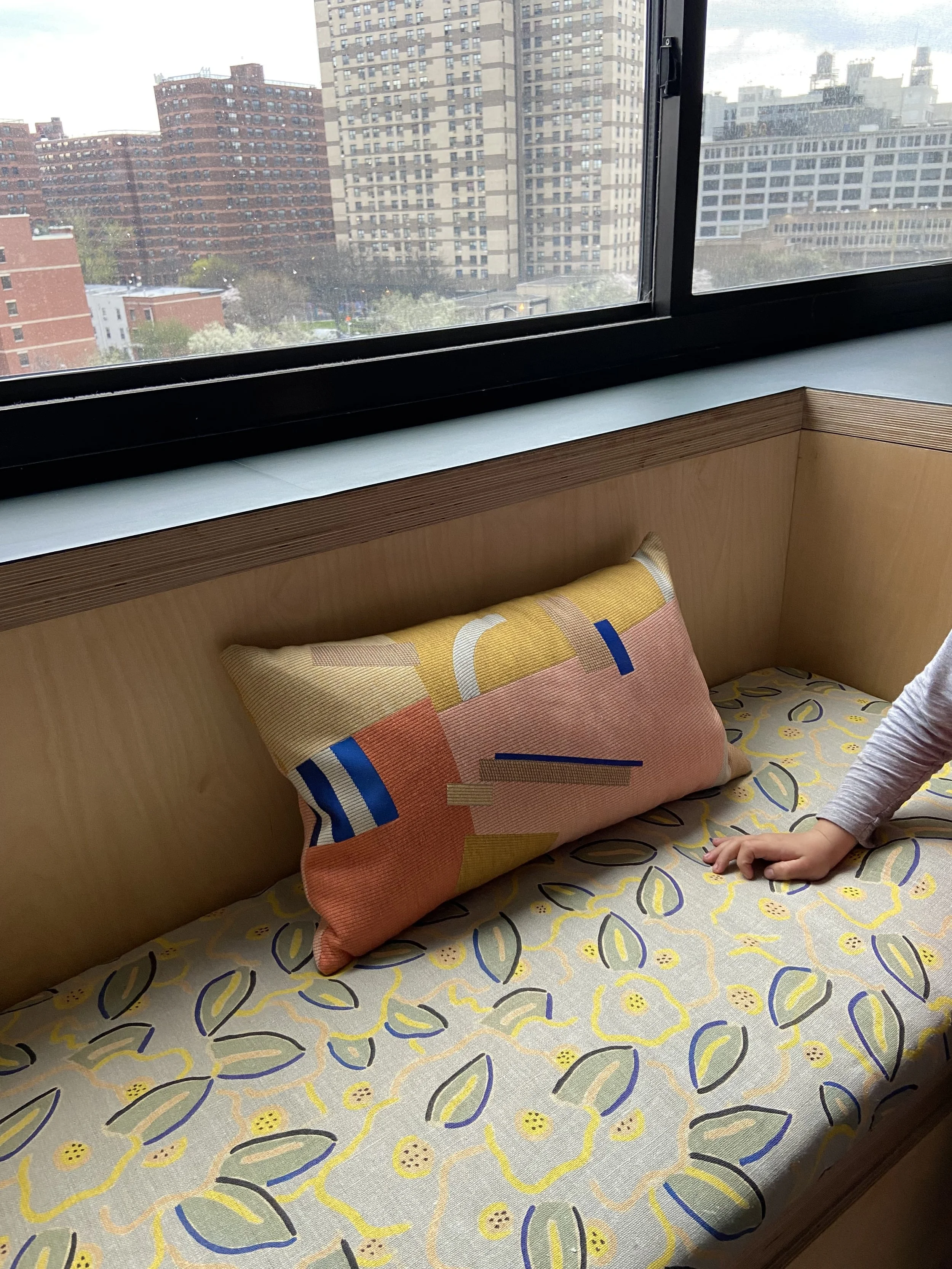 A window seat with a colorful pillow on a floral-patterned cushion, and a child's hand resting on the cushion.