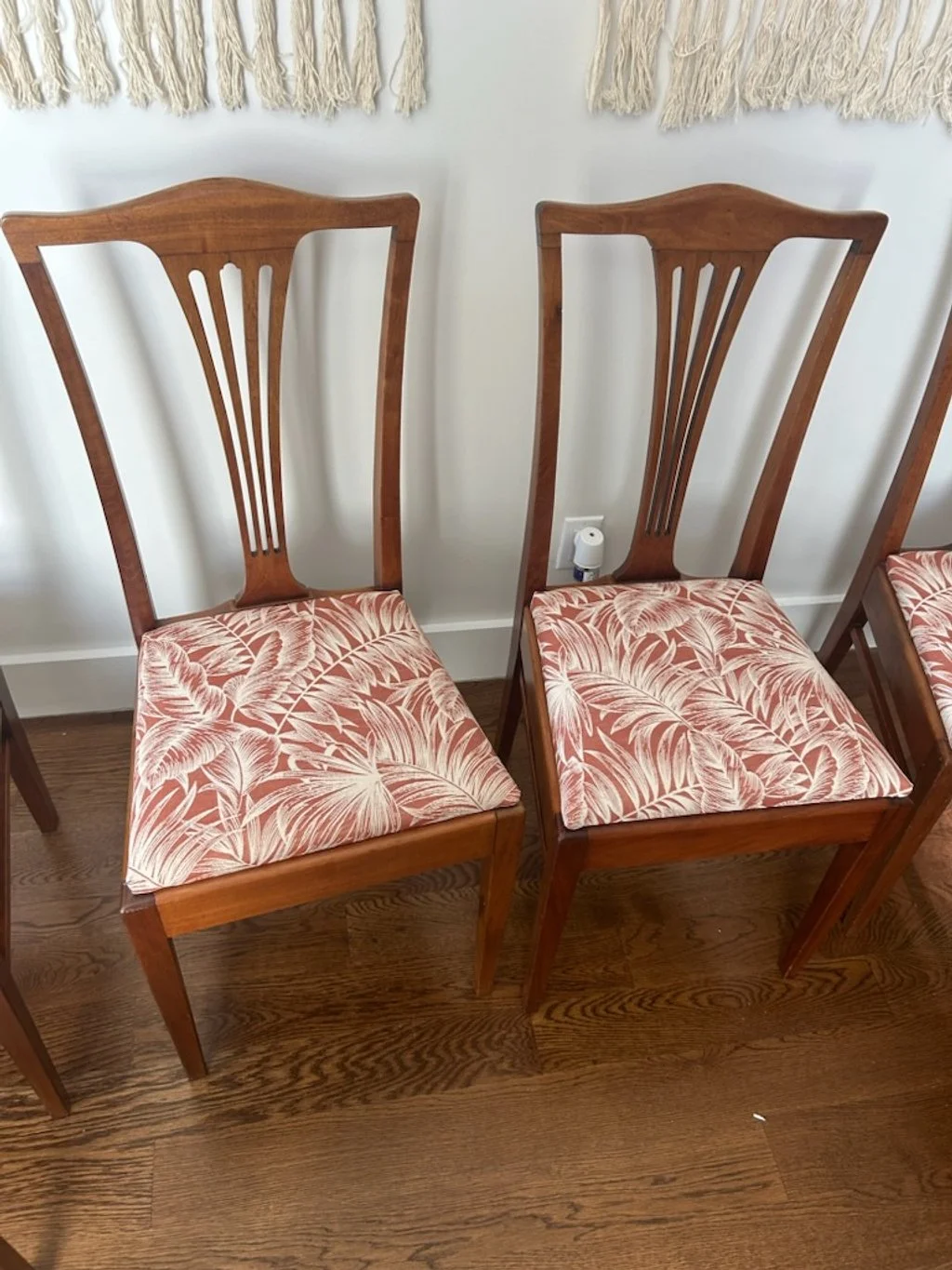 Two wooden dining chairs with tall, curved backs and pink floral cushioned seats, placed on a wooden floor against a white wall with a decorative wall hanging.