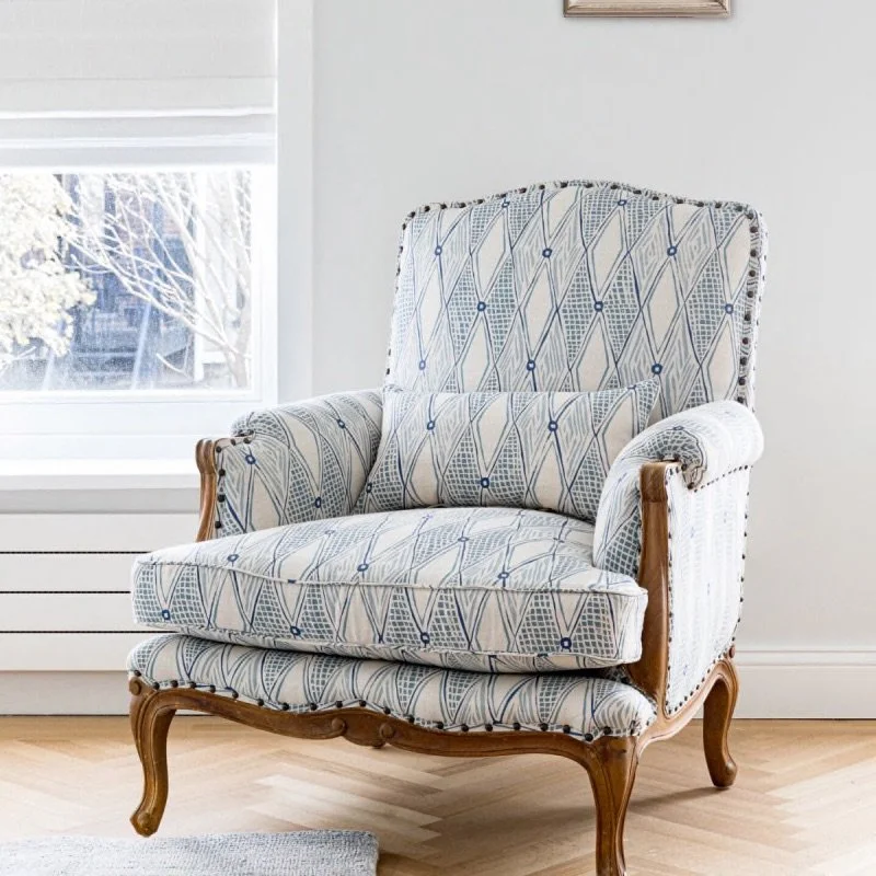 A custom upholstered armchair with a patterned fabric in blue and beige, featuring wooden legs and armrest accents, positioned in a bright room near a window.