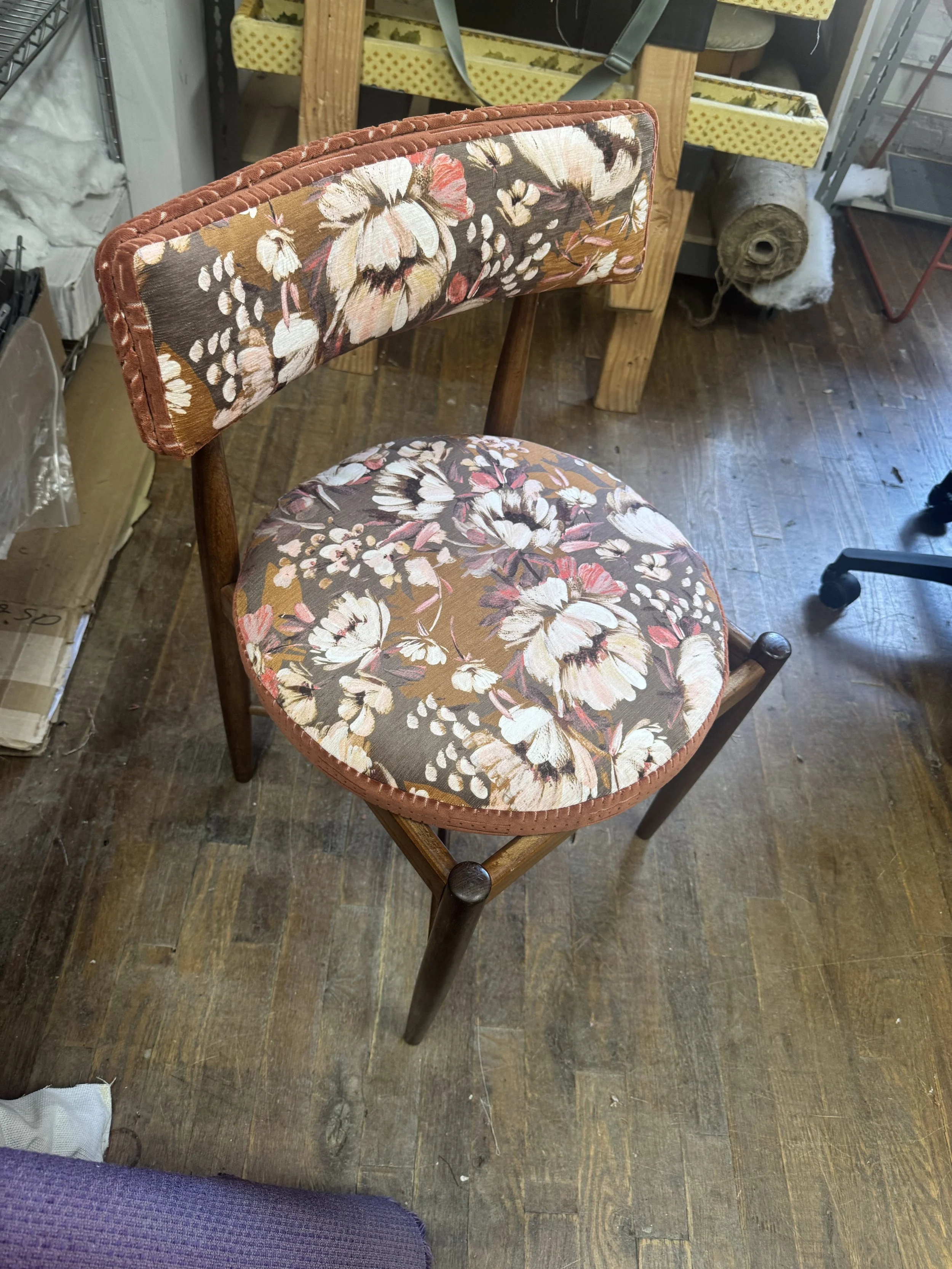 A vintage wooden chair with a floral fabric cushion and backrest, featuring white, pink, and beige flowers on a brown background.