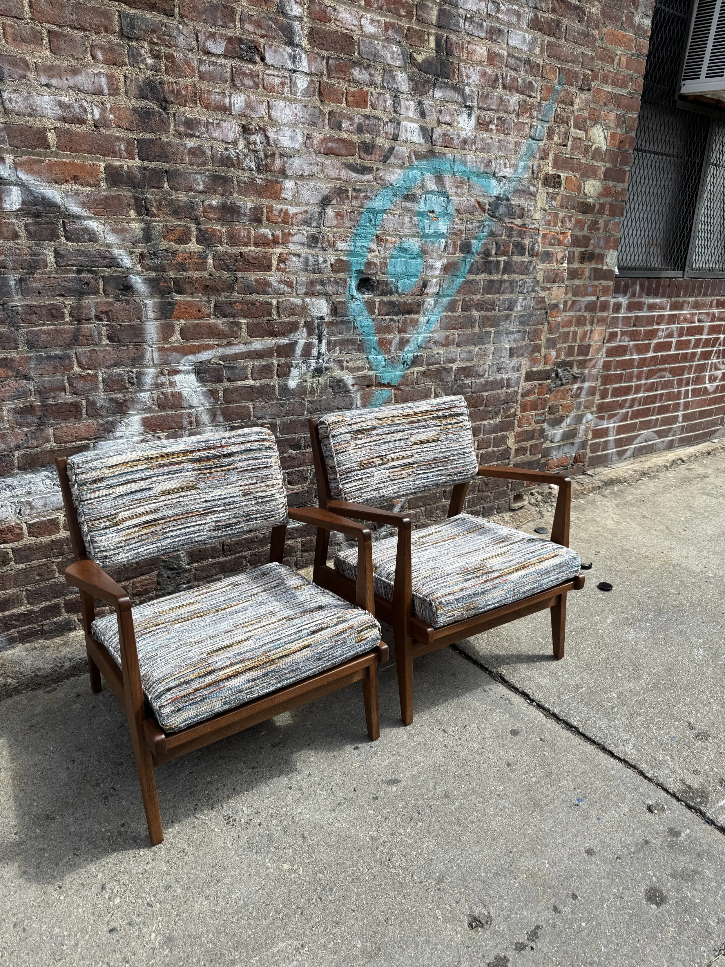 Two vintage wooden armchairs with custom upholstered fabric cushions are placed outdoors against a brick wall with graffiti art.
