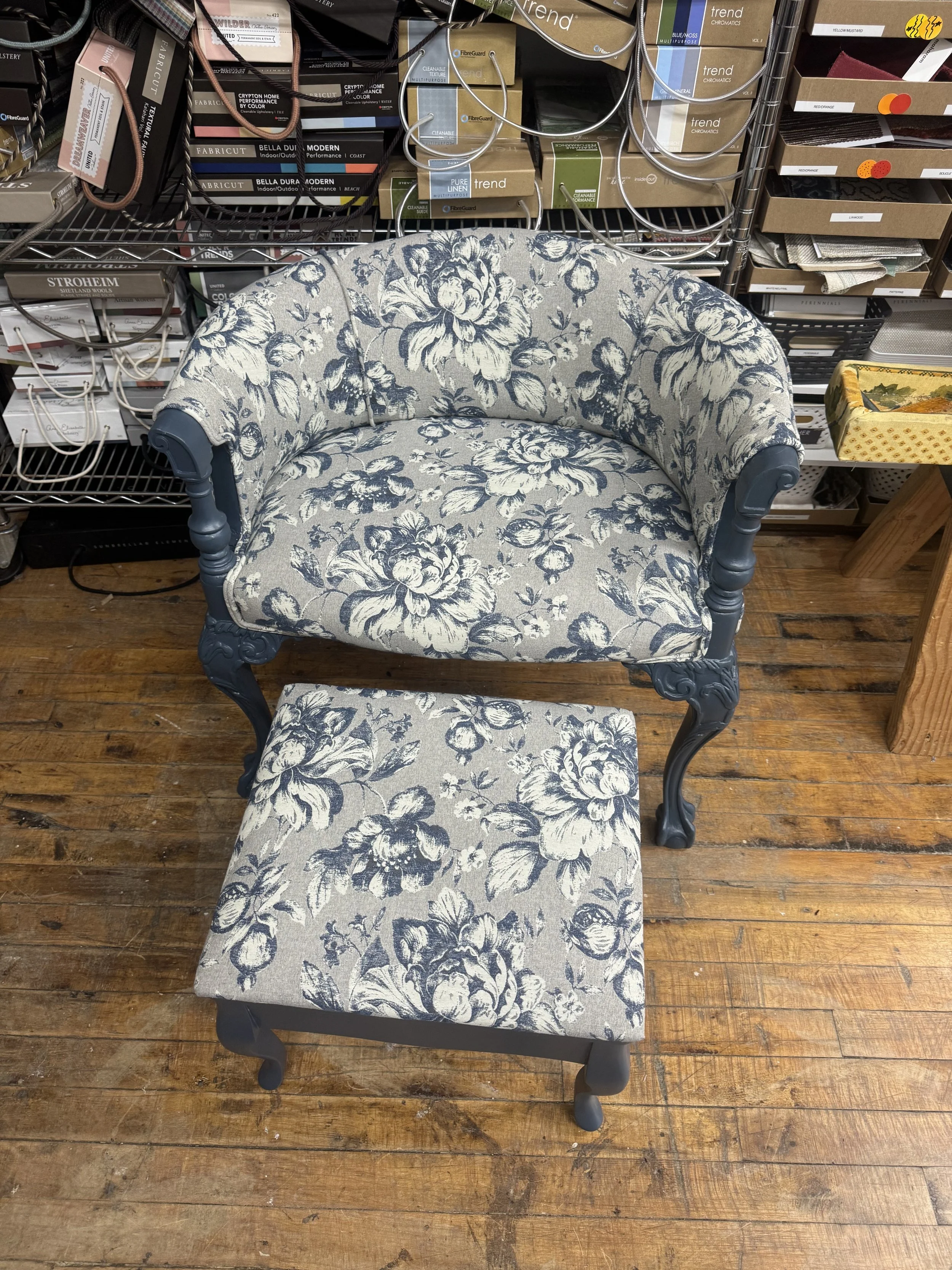 A vintage-style armchair with matching footstool, both upholstered in floral fabric with blue and white pattern, set on a wooden floor in a store.
