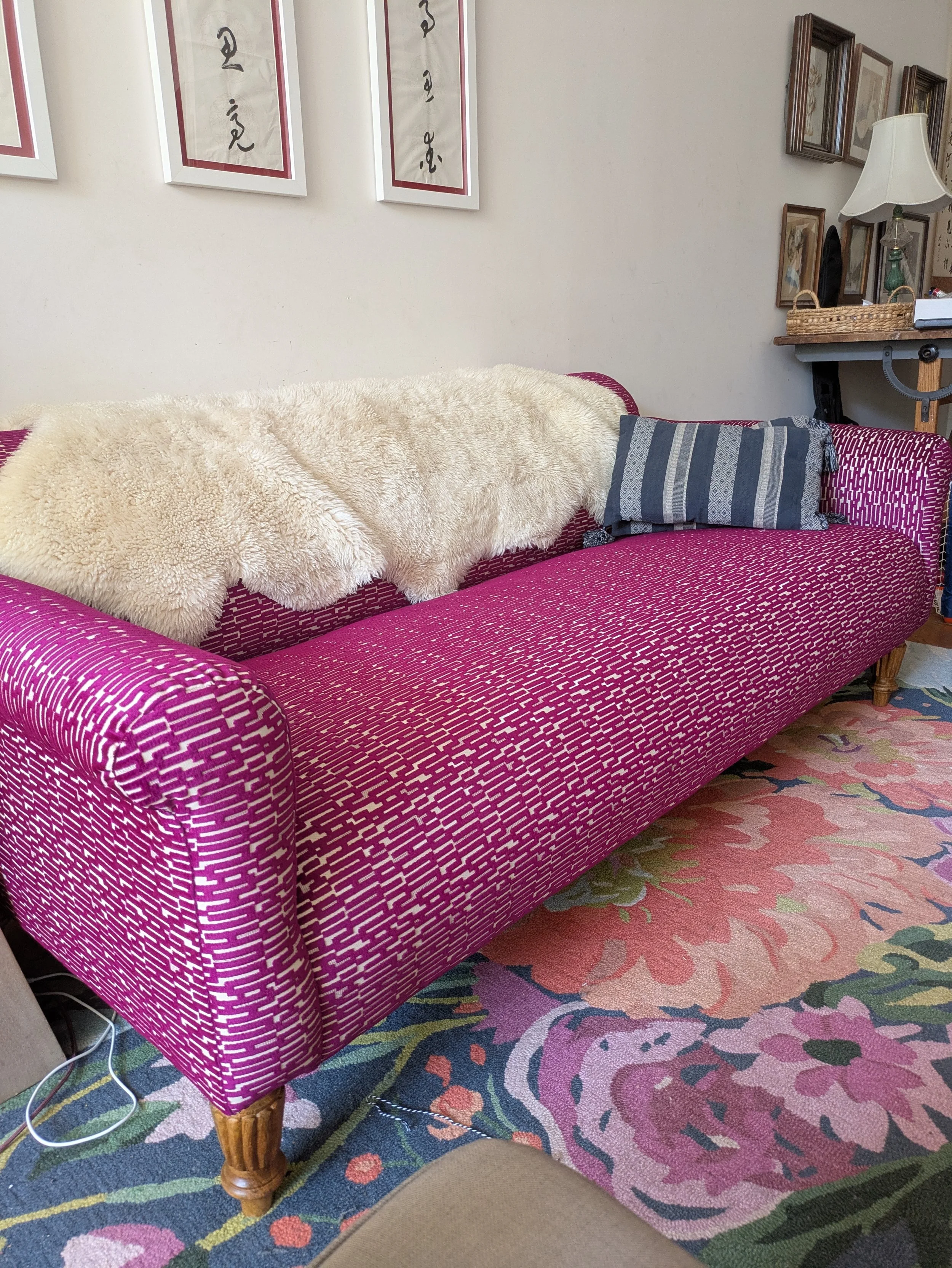 A custom-upholstered pink textured sofa with wooden legs, located in a decorated living room with framed artwork and a floral area rug.
