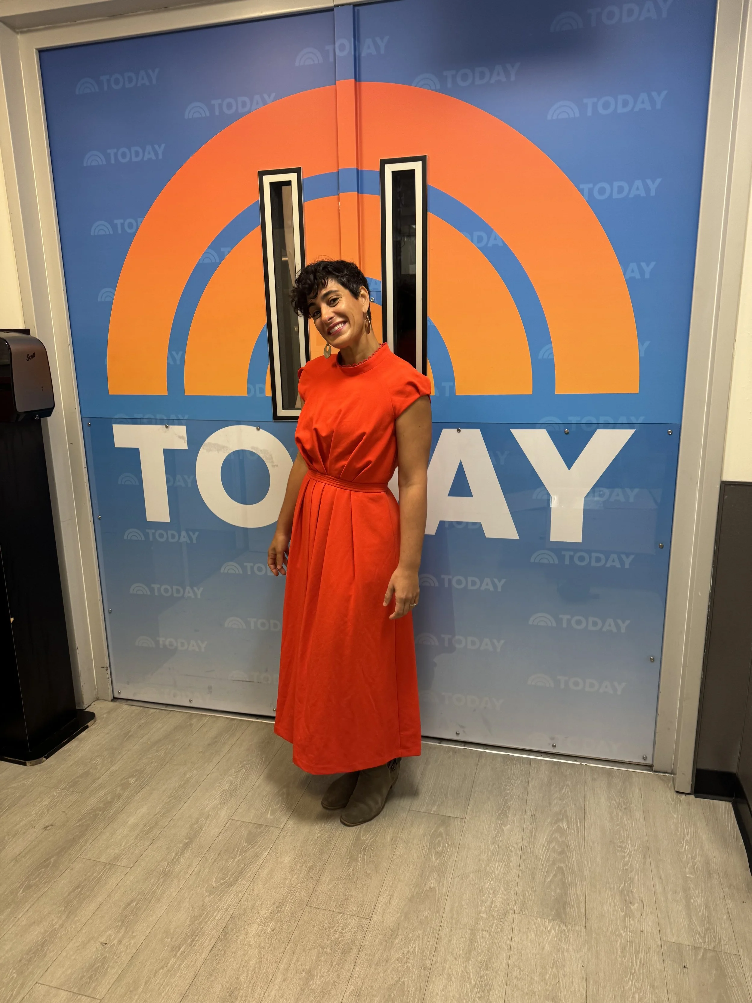 Ruti in a red dress smiling in front of the 'Today' show logo on a blue background.