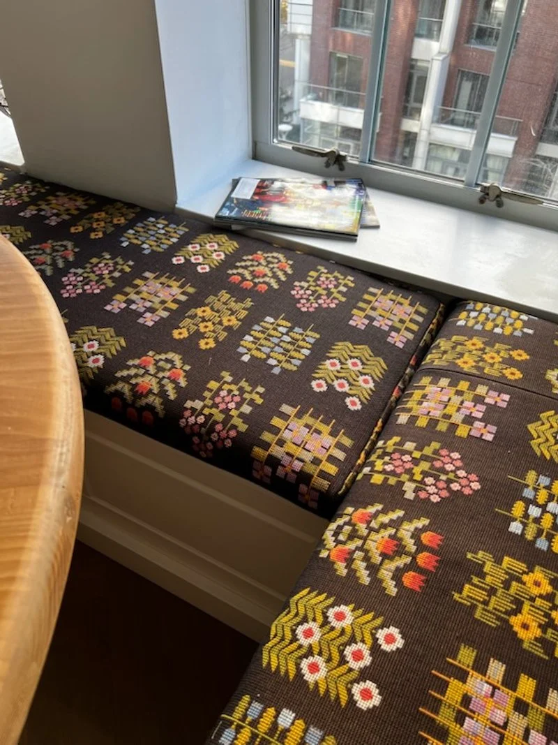 Corner of a window seat with floral patterned cushion, with magazines on the white window sill, overlooking a cityscape.