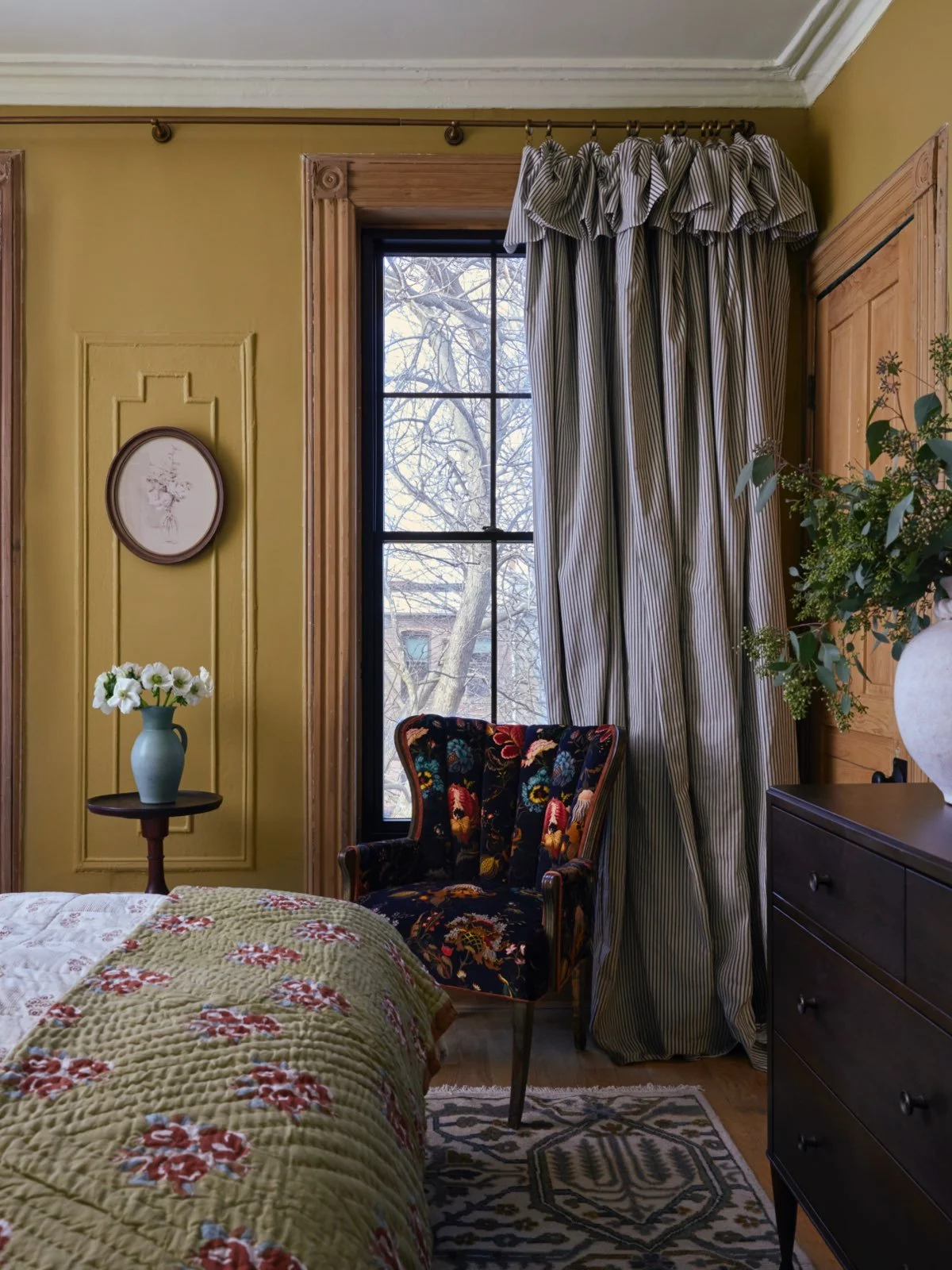 A bedroom corner featuring a custom upholstered floral armchair, sitting against a window with striped curtains.
