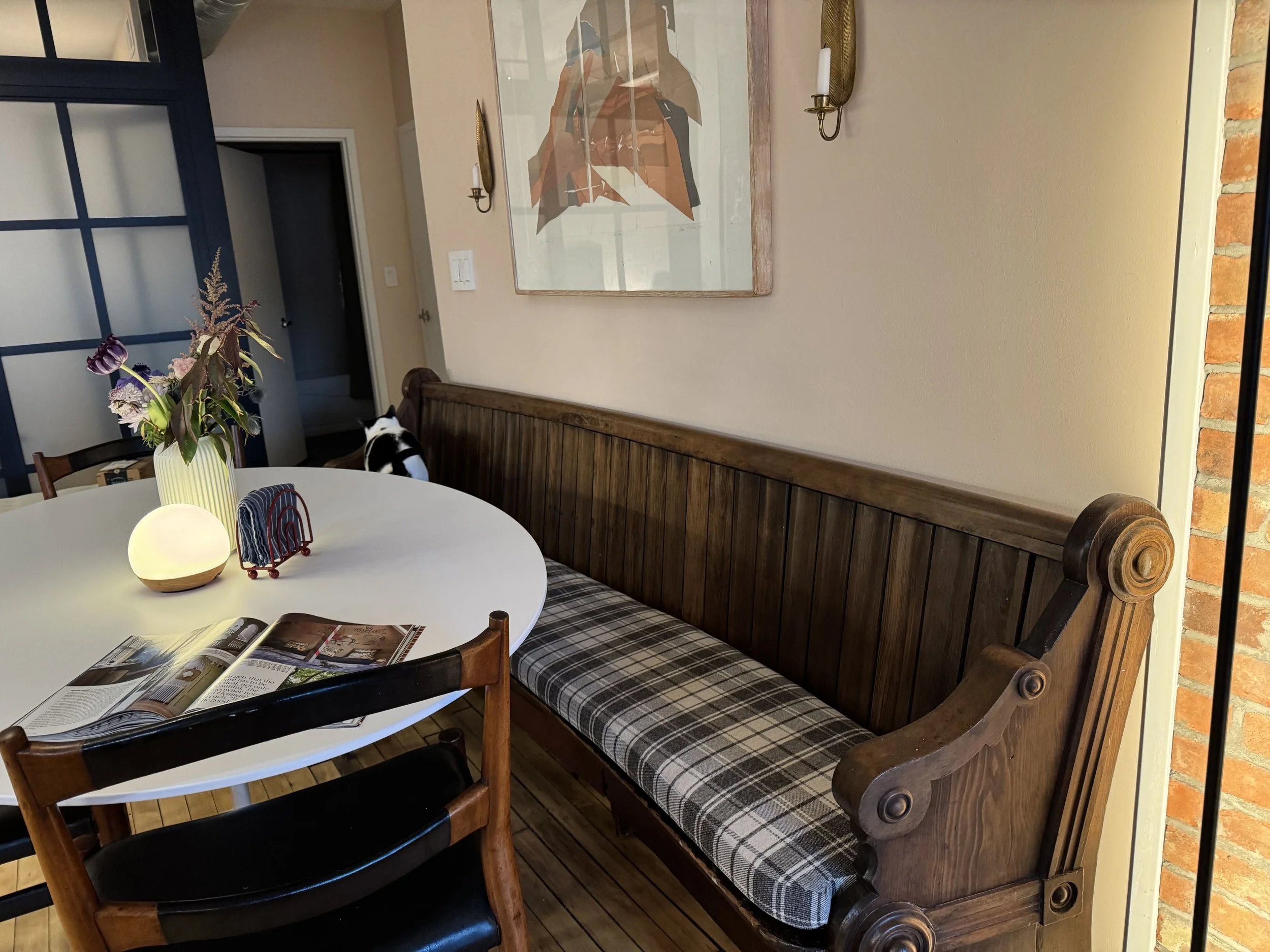 A dining room corner featuring a wooden bench with a custom plaid cushion, a round white table with a vase of flowers, and some accessories.