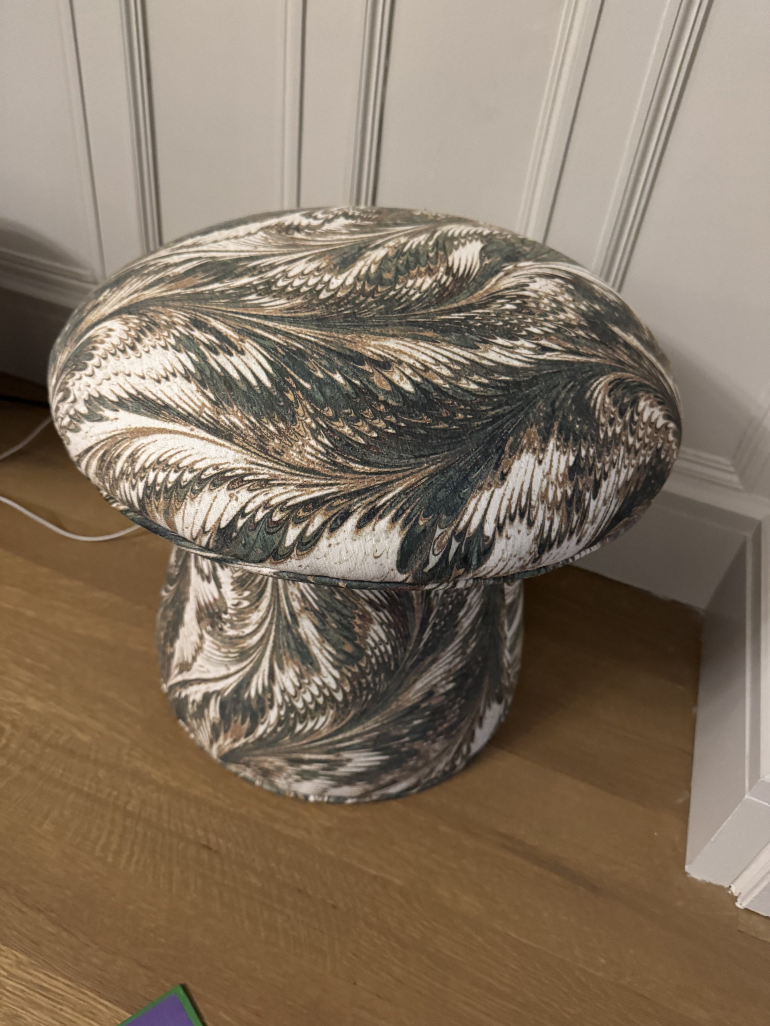 Decorative fabric-covered stool with a feather pattern, placed on wooden floor.