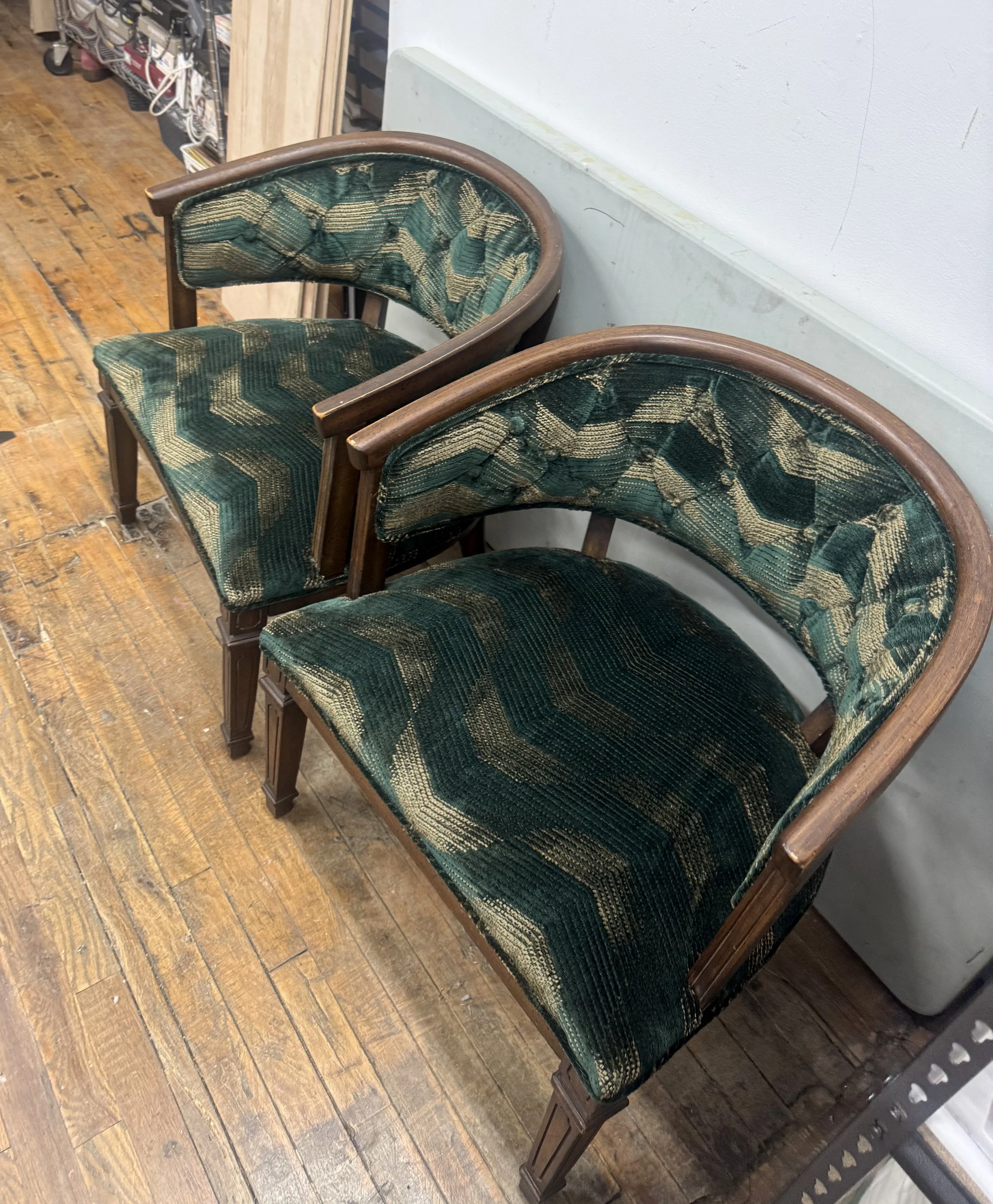 Two vintage green upholstered armchairs with wooden frames, placed side by side on a wooden floor against a white wall.