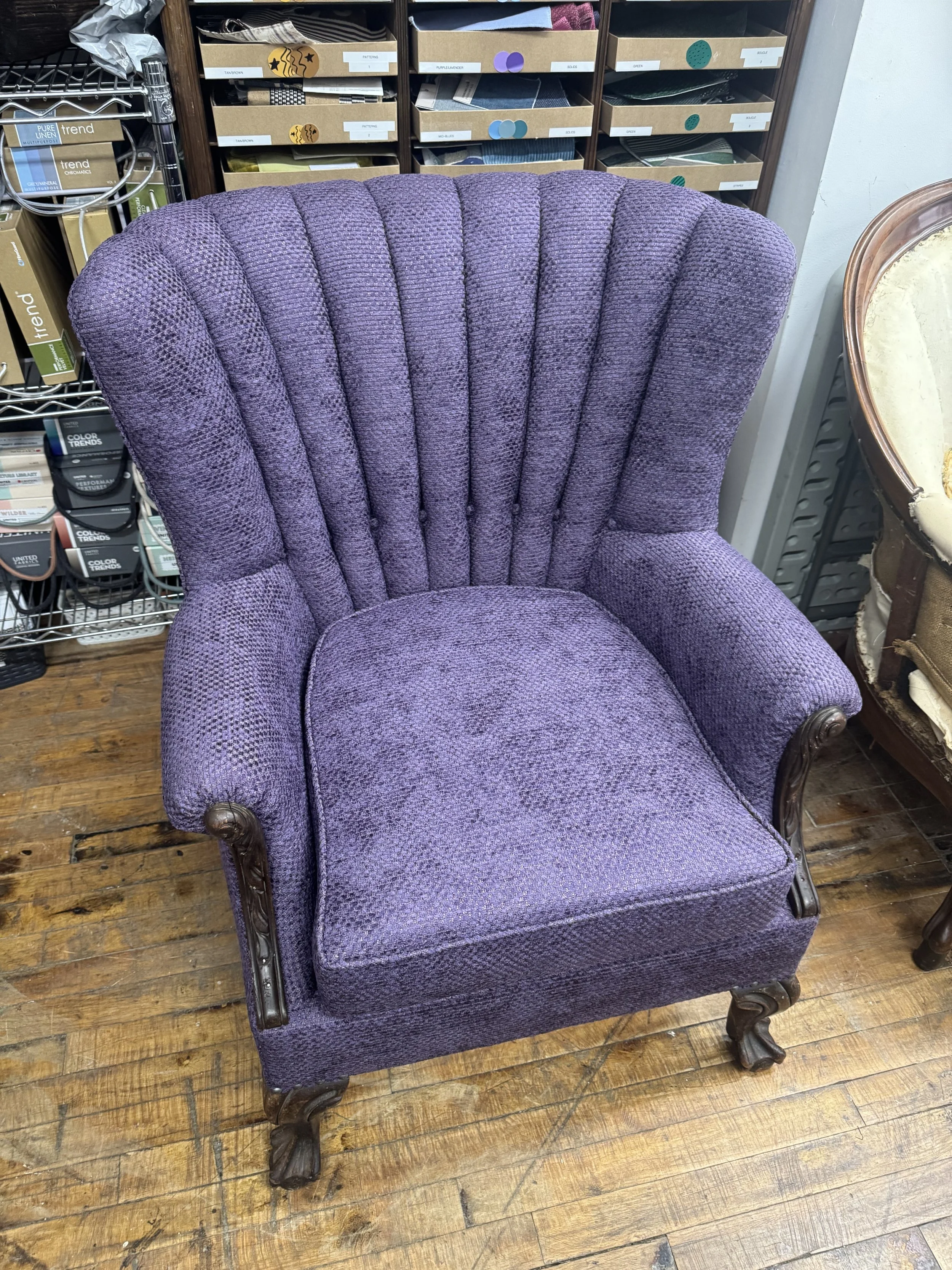 Vintage purple upholstered armchair with dark wooden carved legs and arm accents, set on a wooden floor in the workshop.