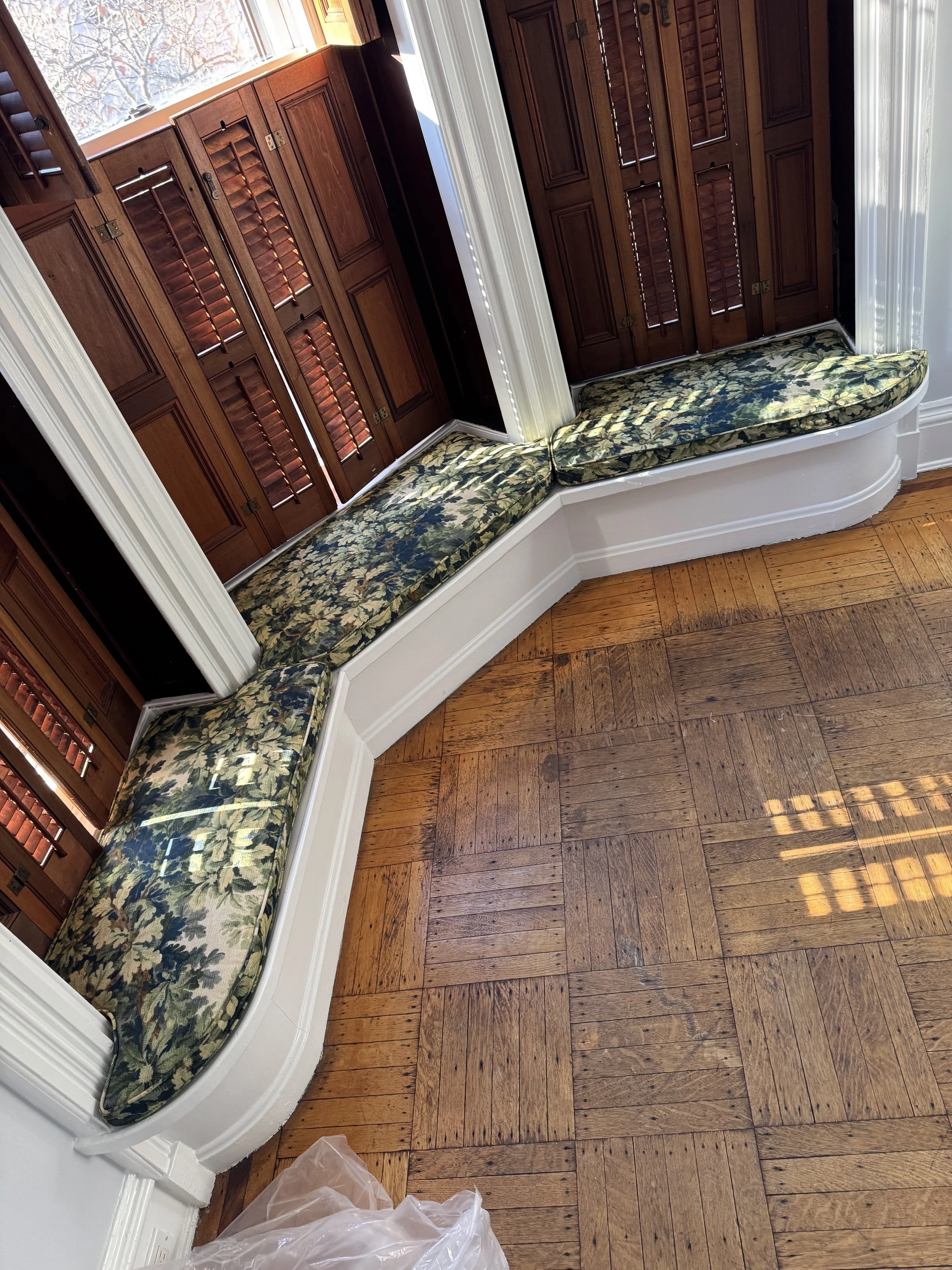 A corner window seat with a floral cushion in front of three wooden shuttered windows, in a room with wooden flooring and white molding.