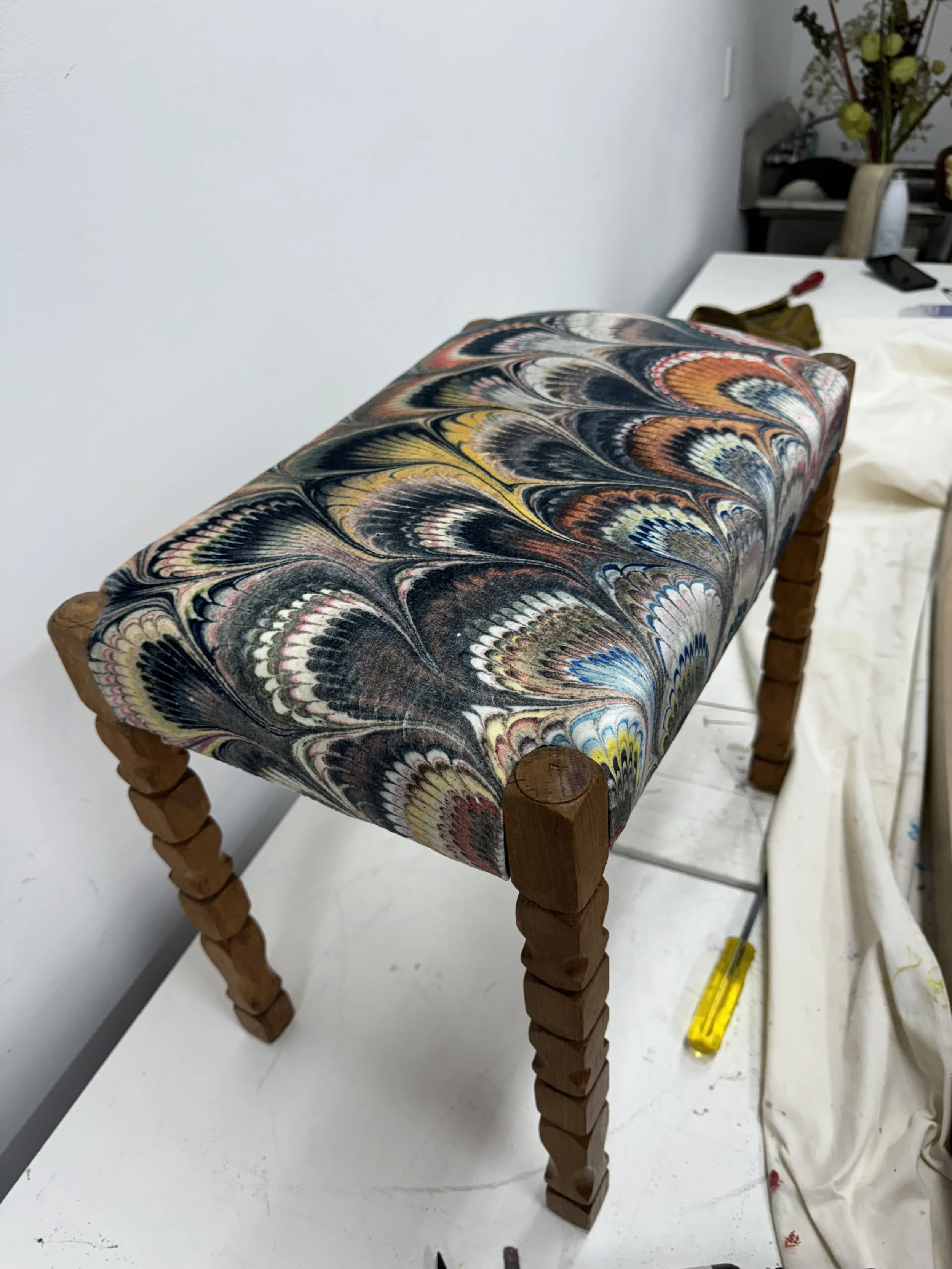 A wooden bench with a cushion featuring a colorful marbled pattern in black, yellow, orange, blue, and white, positioned on a work surface with tools and fabric.
