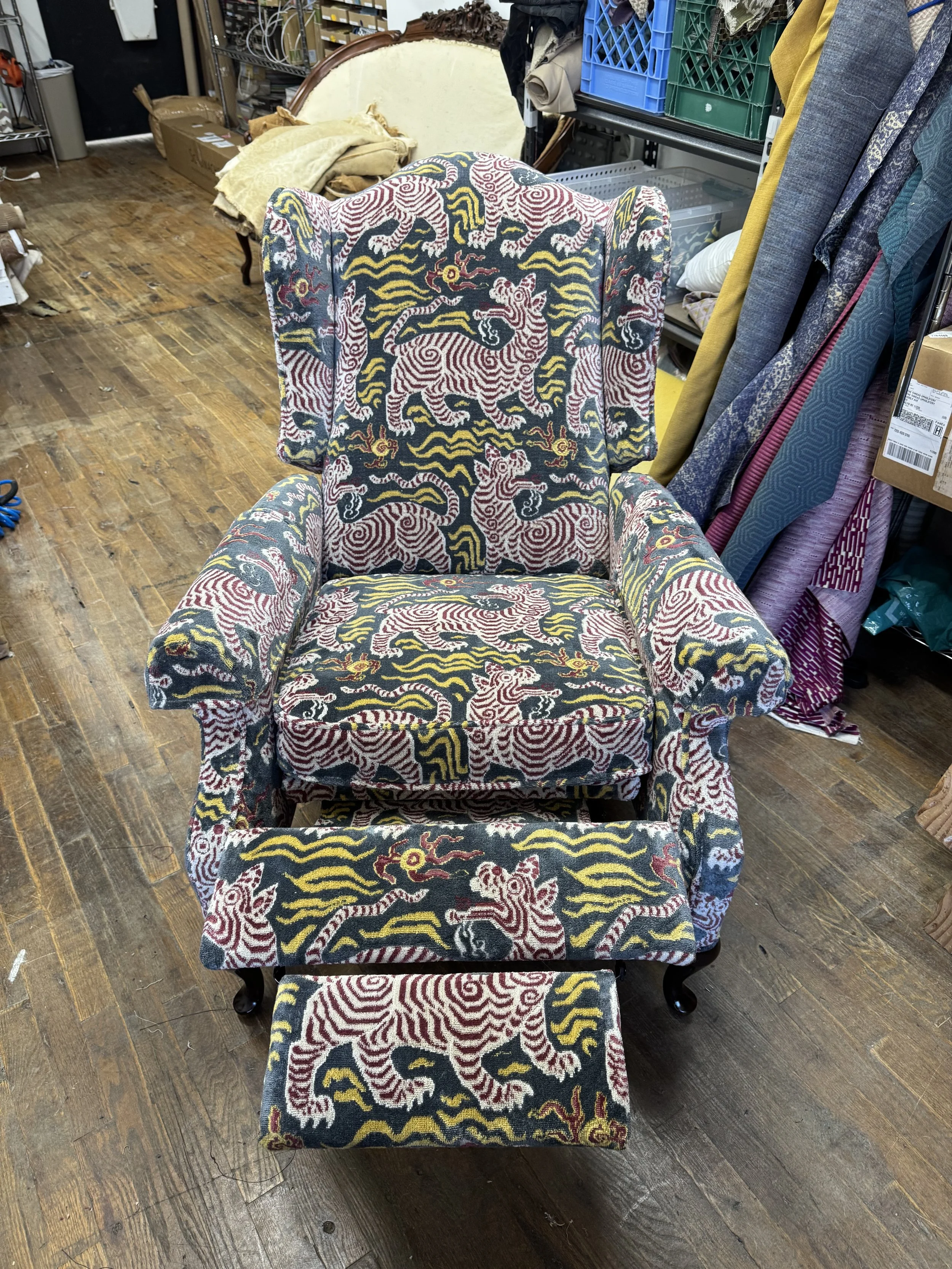 A vintage-style armchair with a matching ottoman, covered in fabric featuring a pattern of stylized tigers and swirling designs in red, yellow, and white on a dark background, in Ruti's workshop.