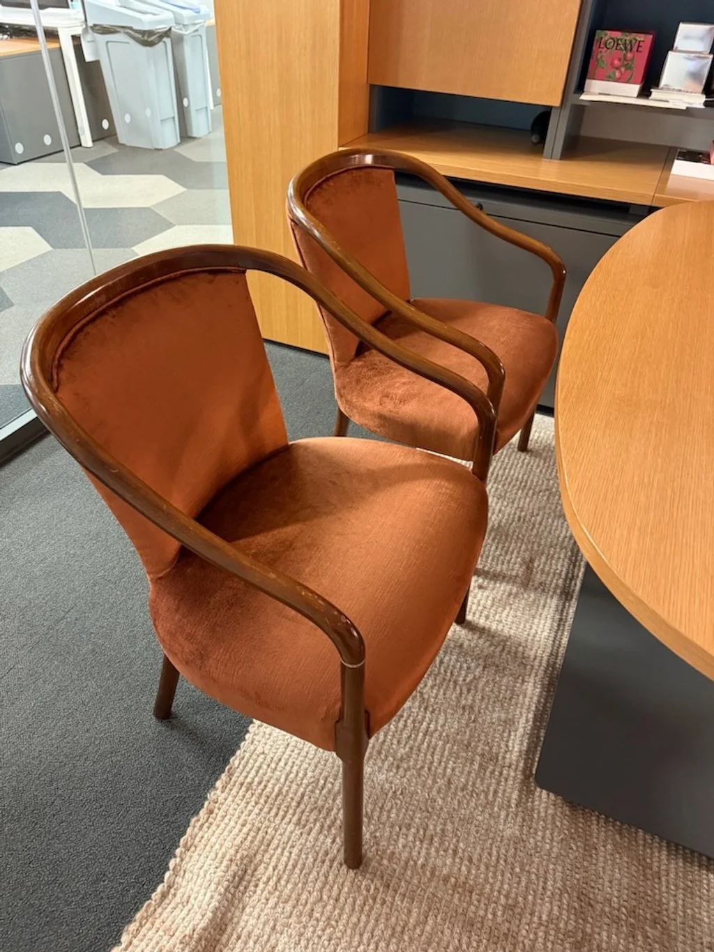 Two vintage orange upholstered chairs with wooden armrests and legs are positioned next to a round wooden table in an office.