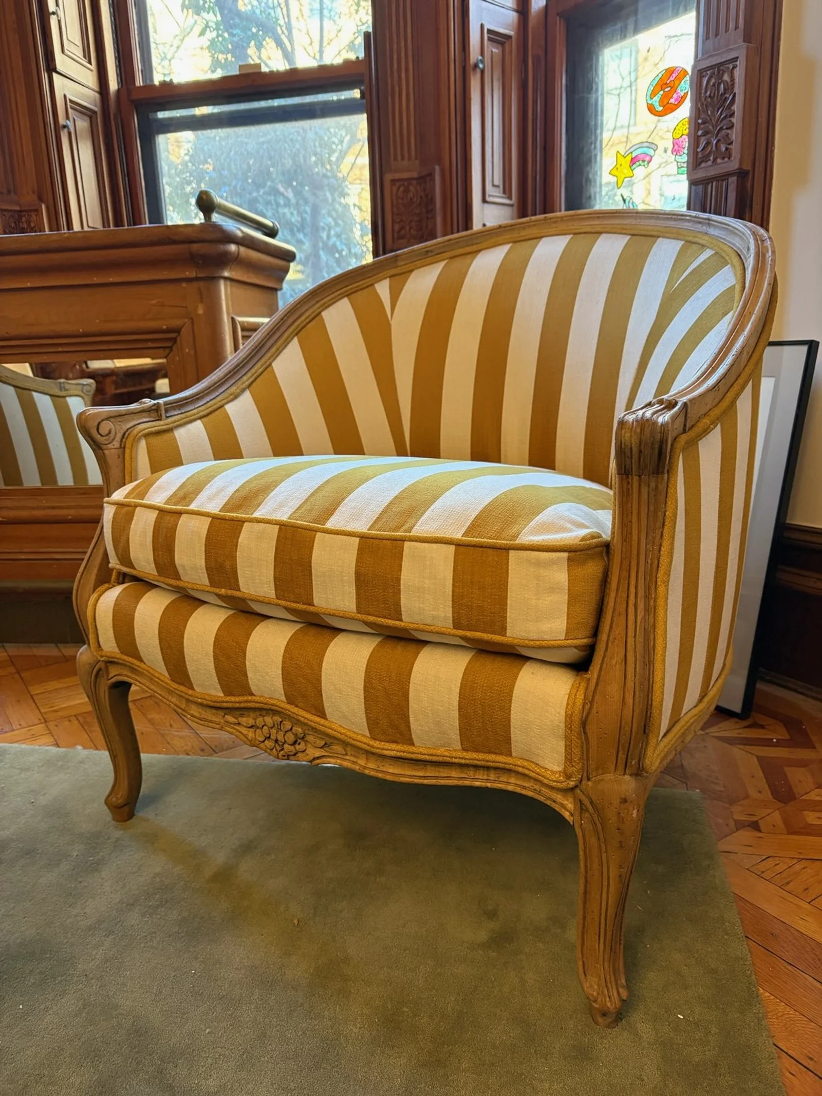 A vintage-style wooden sofa with yellow and white striped upholstery, placed on a carpeted floor in a room with large windows.