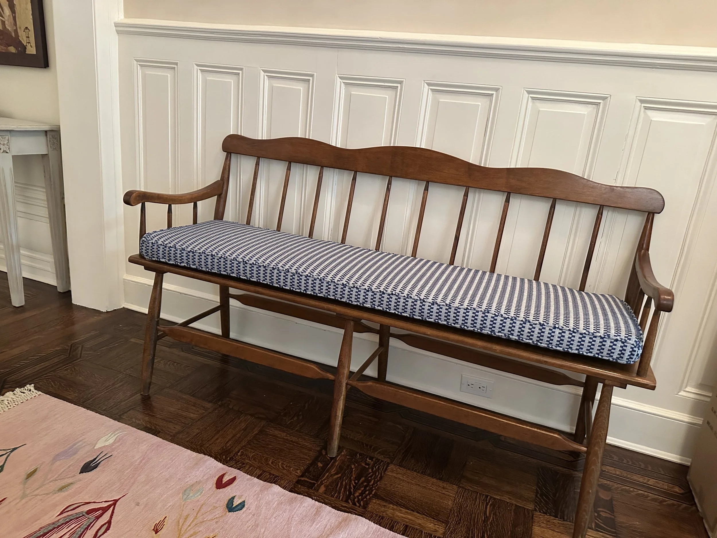 Wooden bench with blue and white patterned cushion in a room with wood flooring and white paneled wall.