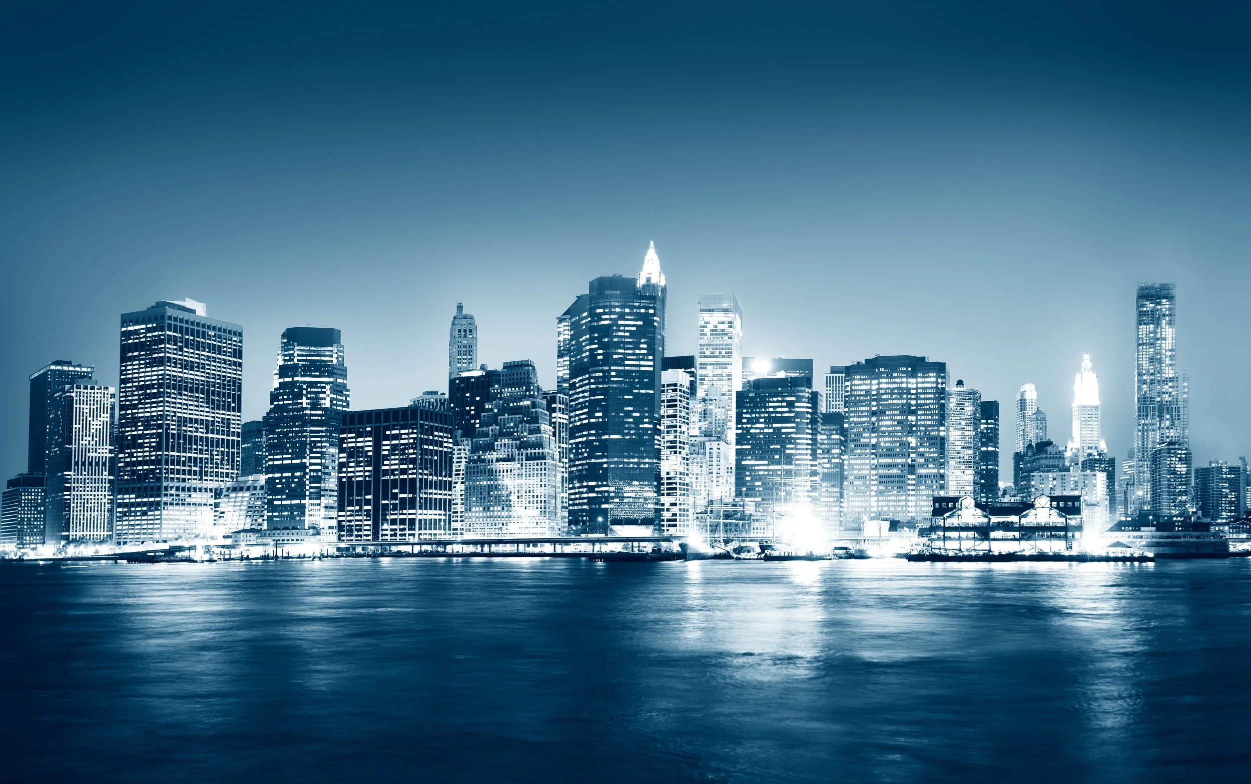 Nighttime city skyline with illuminated skyscrapers and reflections on the water.