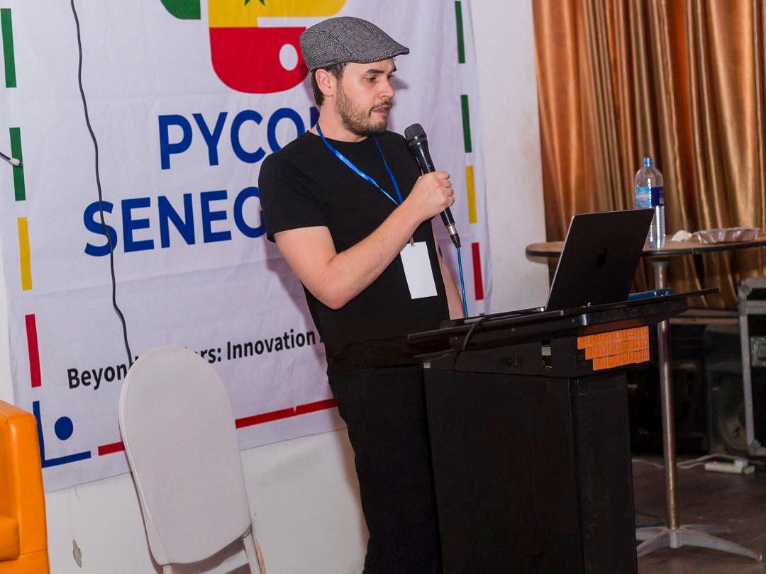 A man with a gray flat cap and black t-shirt speaking into a microphone while standing at a lectern with a laptop. Behind him is a colorful banner with blue, red, yellow, and green text that reads 'PYCON SENEGA' and part of a phrase 'Beyond Tech: Innovation.' A white chair and a table with a water bottle are visible next to him.
