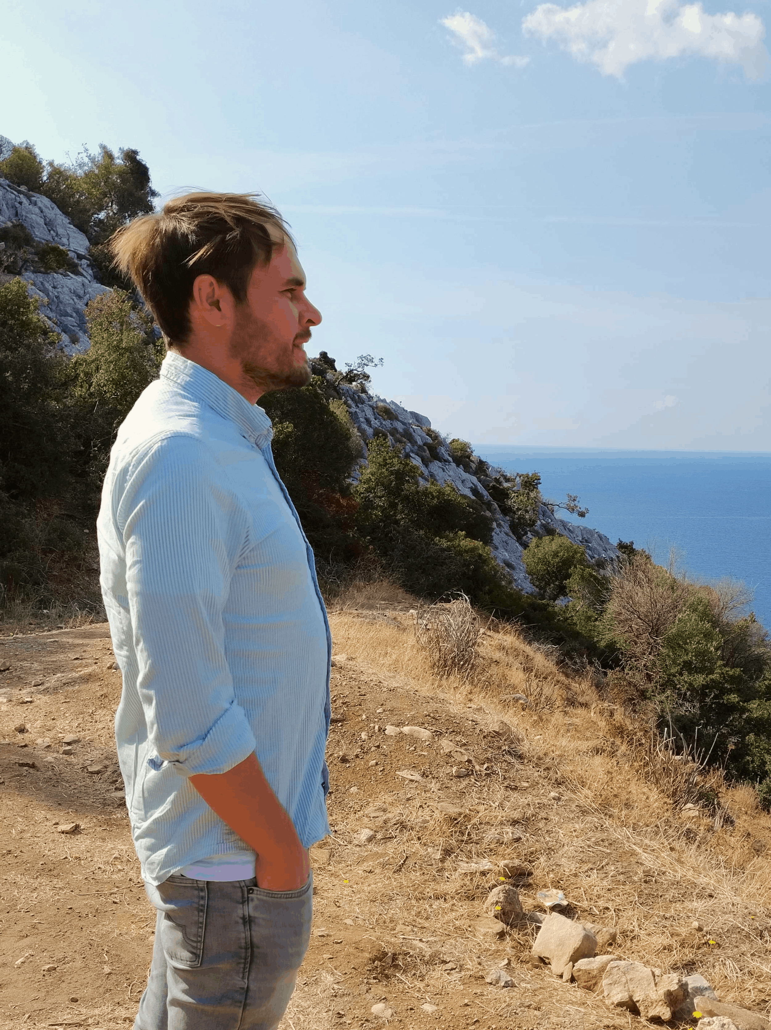 Niek standing on a dirt trail overlooking the ocean with rocky hillside and trees in the background.