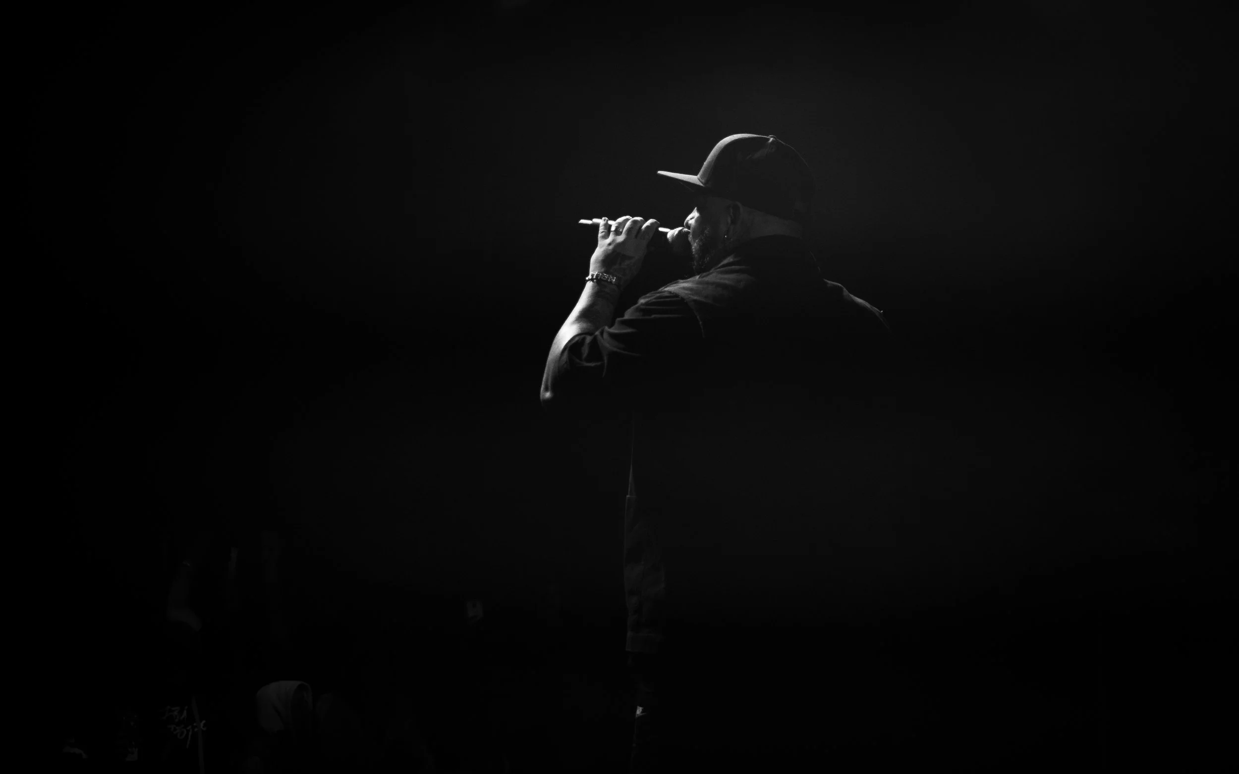 A black and white photo of a man singing into a microphone on stage, wearing a baseball cap and casual clothing, illuminated by a single light source from above.