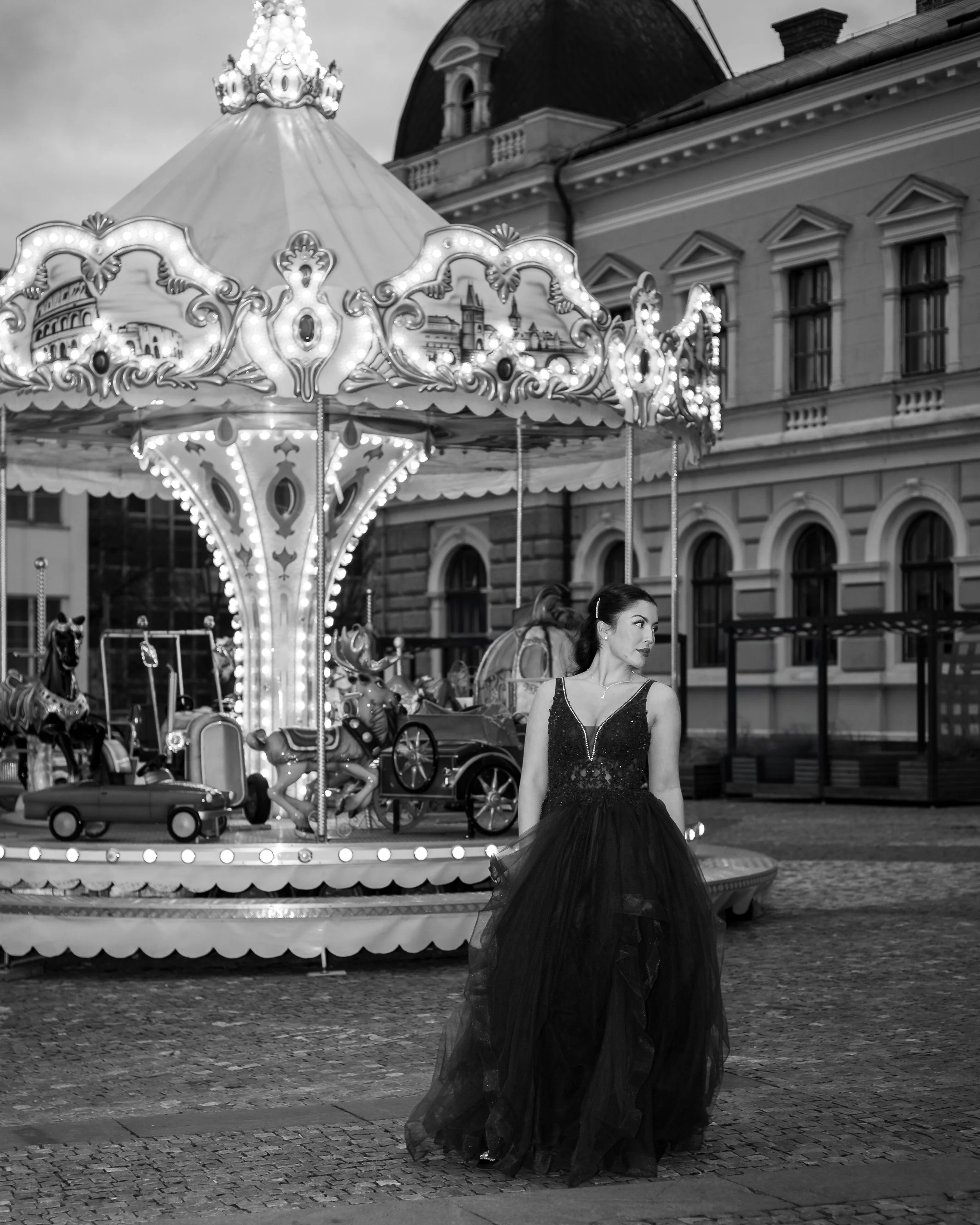 A young woman dressed in a black evening gown with a tulle skirt, standing near an illuminated carousel at an outdoor amusement park. The background features historic buildings with arched windows and ornate architectural details, captured in black and white.