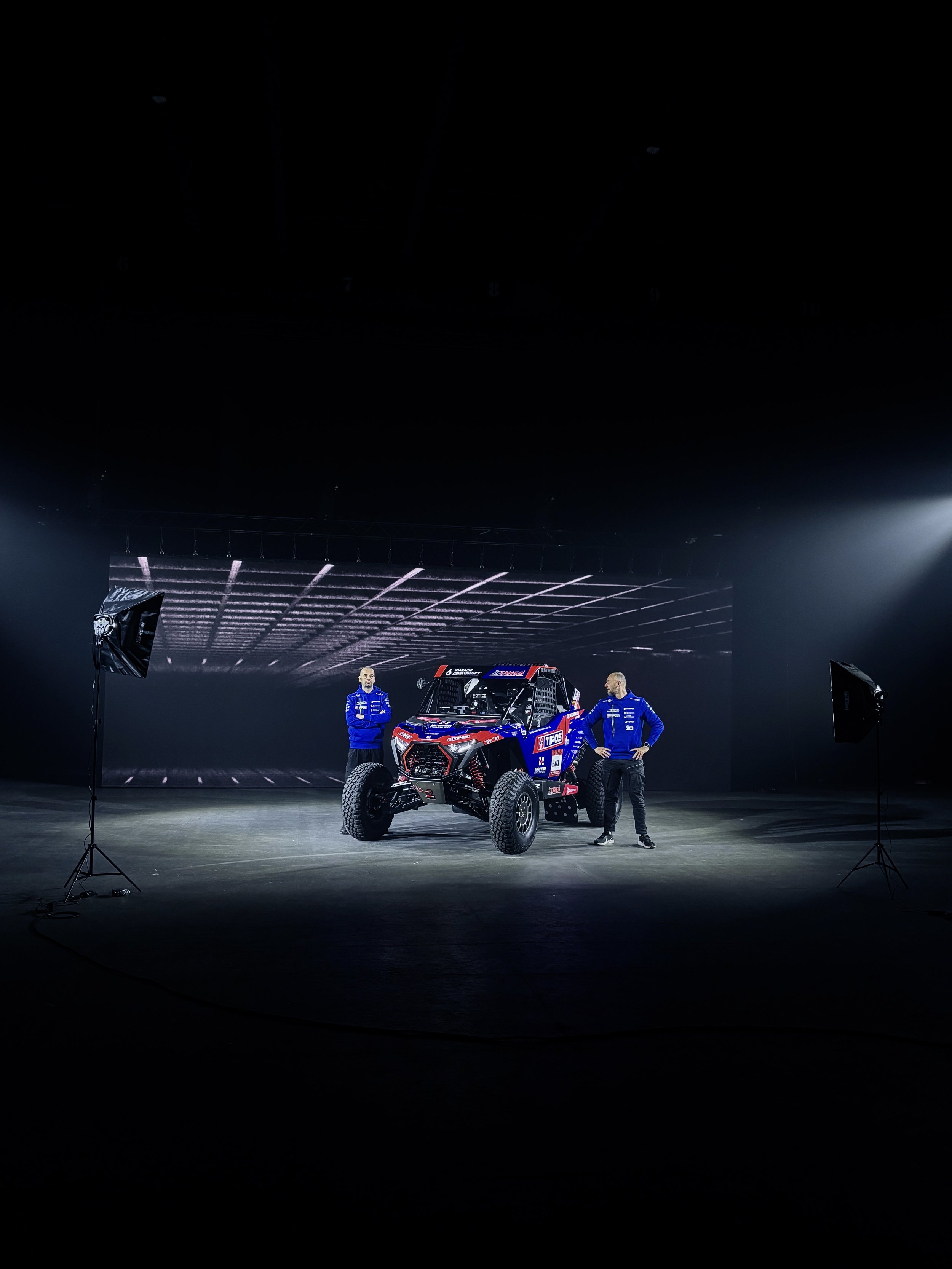 Two men in blue racing suits standing next to a rally car inside a photo studio with a large screen behind them.