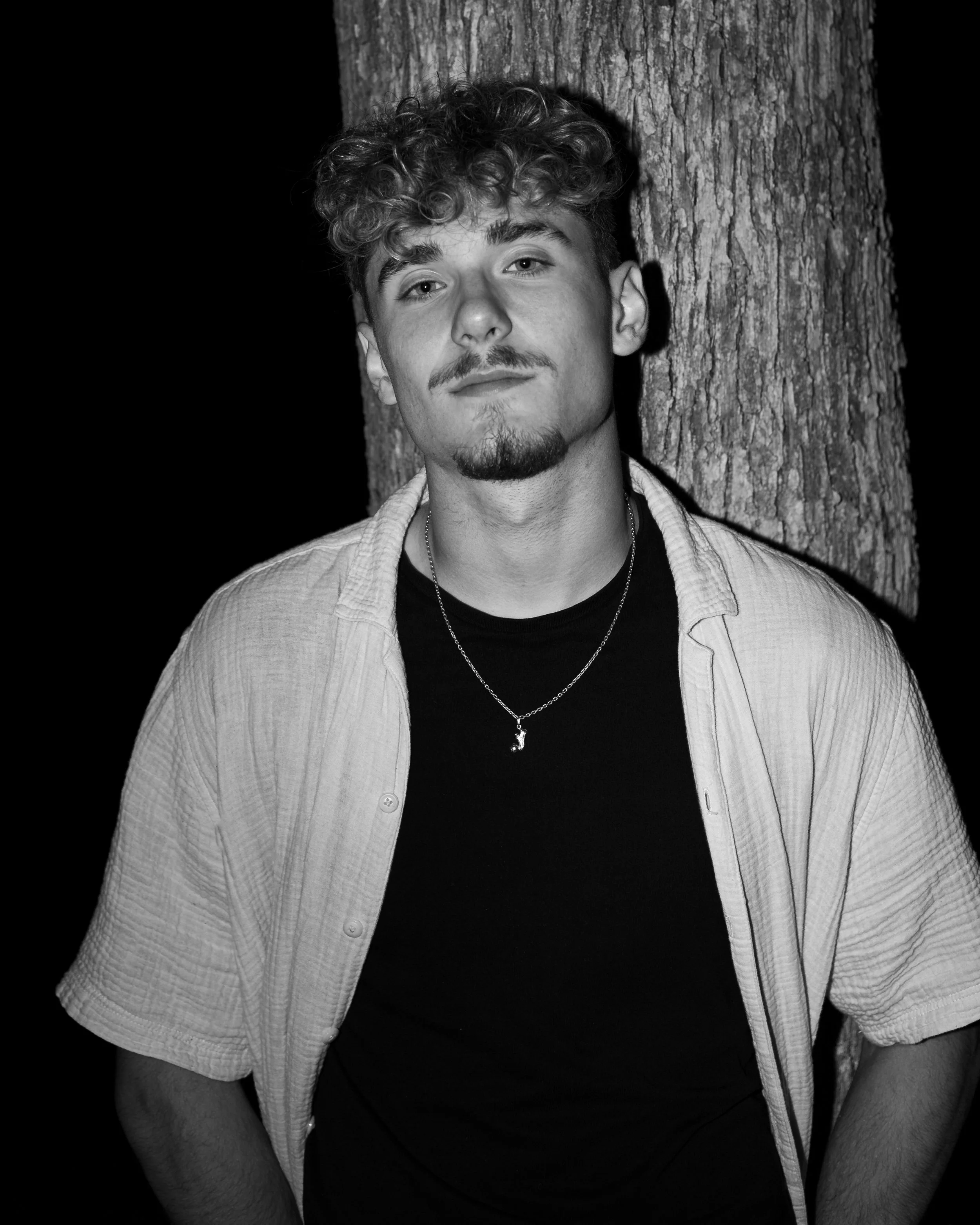 Black and white photo of a young man with curly hair, a goatee, and a slight smile, standing outdoors against a tree trunk at night.