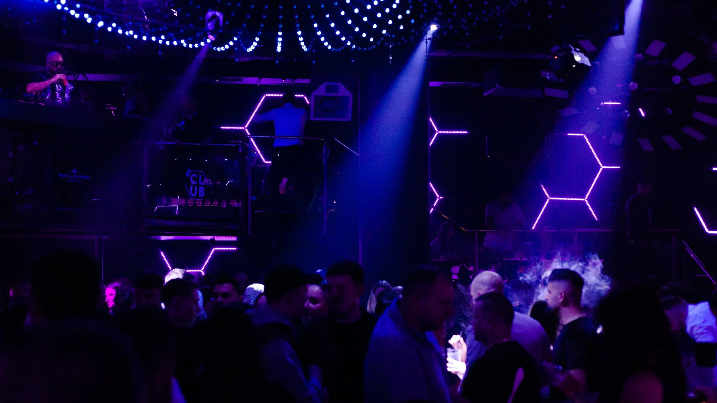 Nightclub scene with purple neon lights, a crowd of people dancing and socializing, and a DJ performing on stage under decorative string lights.