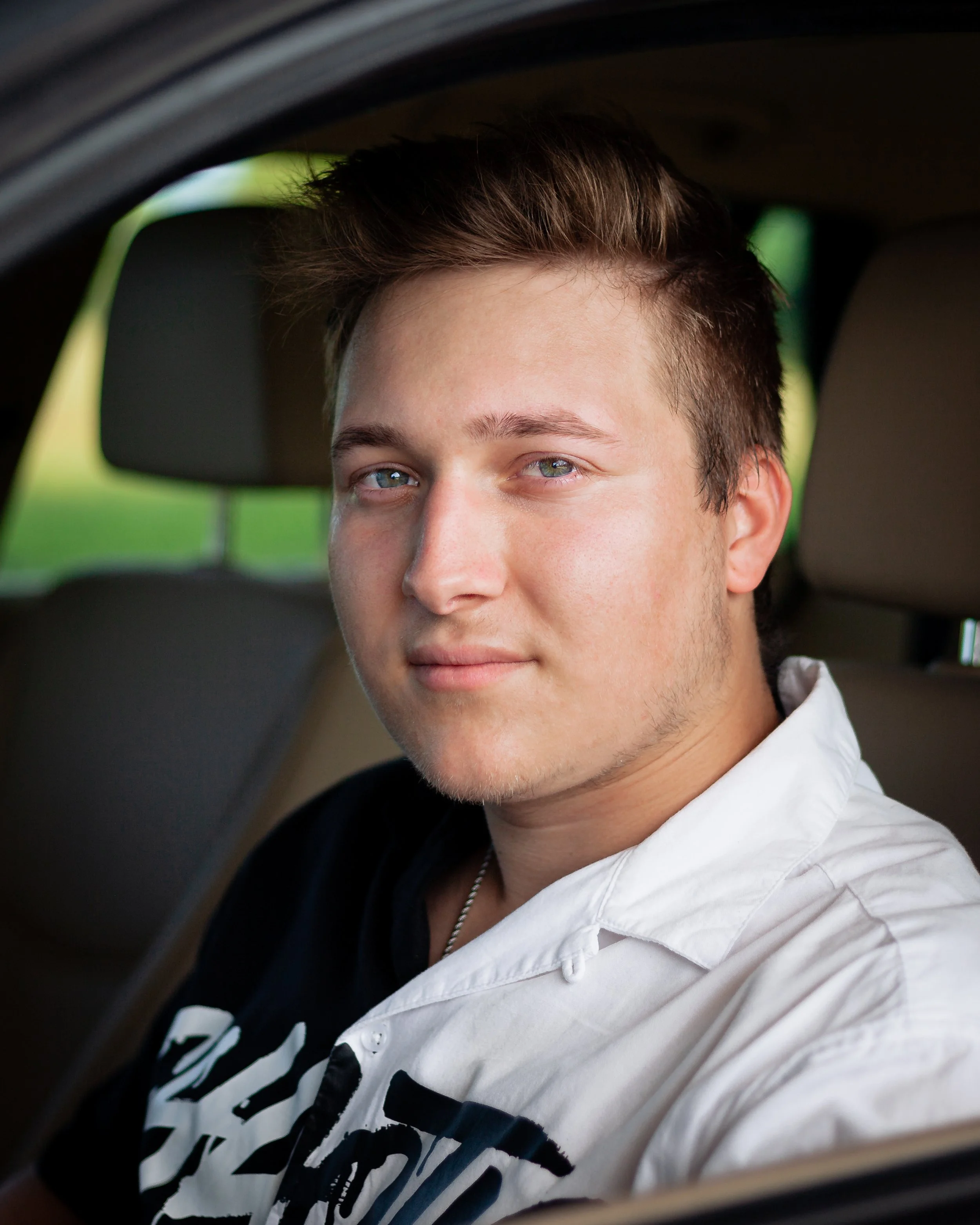 Close-up of a young man's face with light eyes and short hair, sitting in a vehicle with a blurred background.