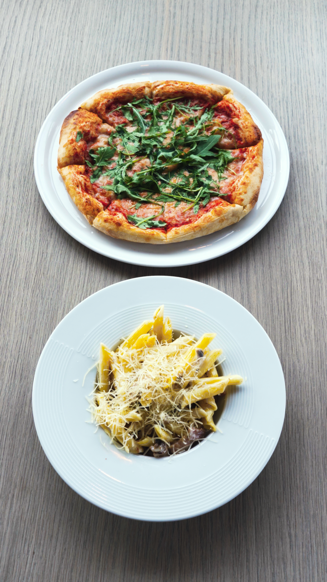 A pepperoni pizza topped with arugula on a white plate and a bowl of penne pasta with grated cheese on a white plate, both on a gray wooden table.